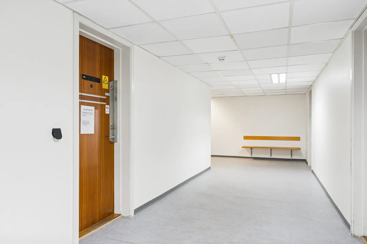 En tom korridor med hvide vægge og lofter og en brun dør med adgangskode. Der er et gult skilt og et hvidt papir med tekst på døren. En smal bænk står mod væggen