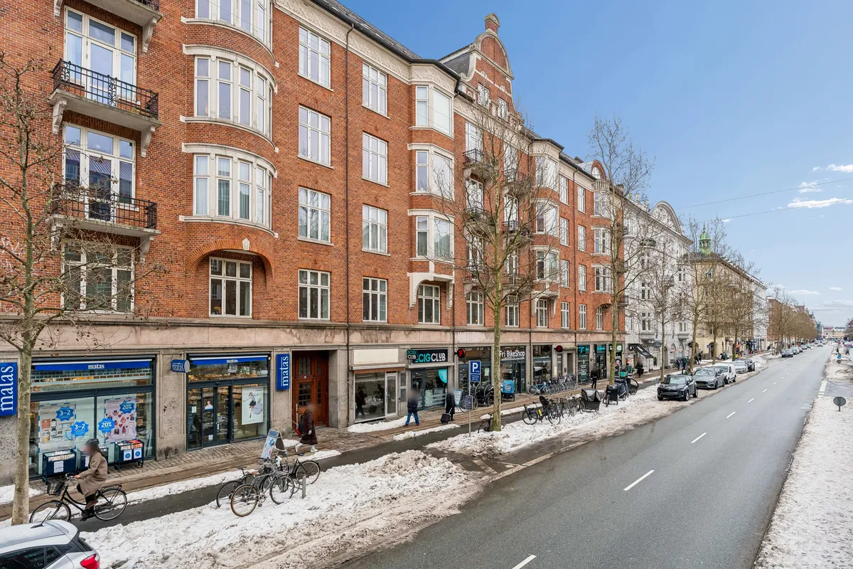 Mennesker kører på cykler og går på sne på Østerbrogade. Mange cykler står parkeret. Butikker er åbne med skilte. Det er sne dekket i området.