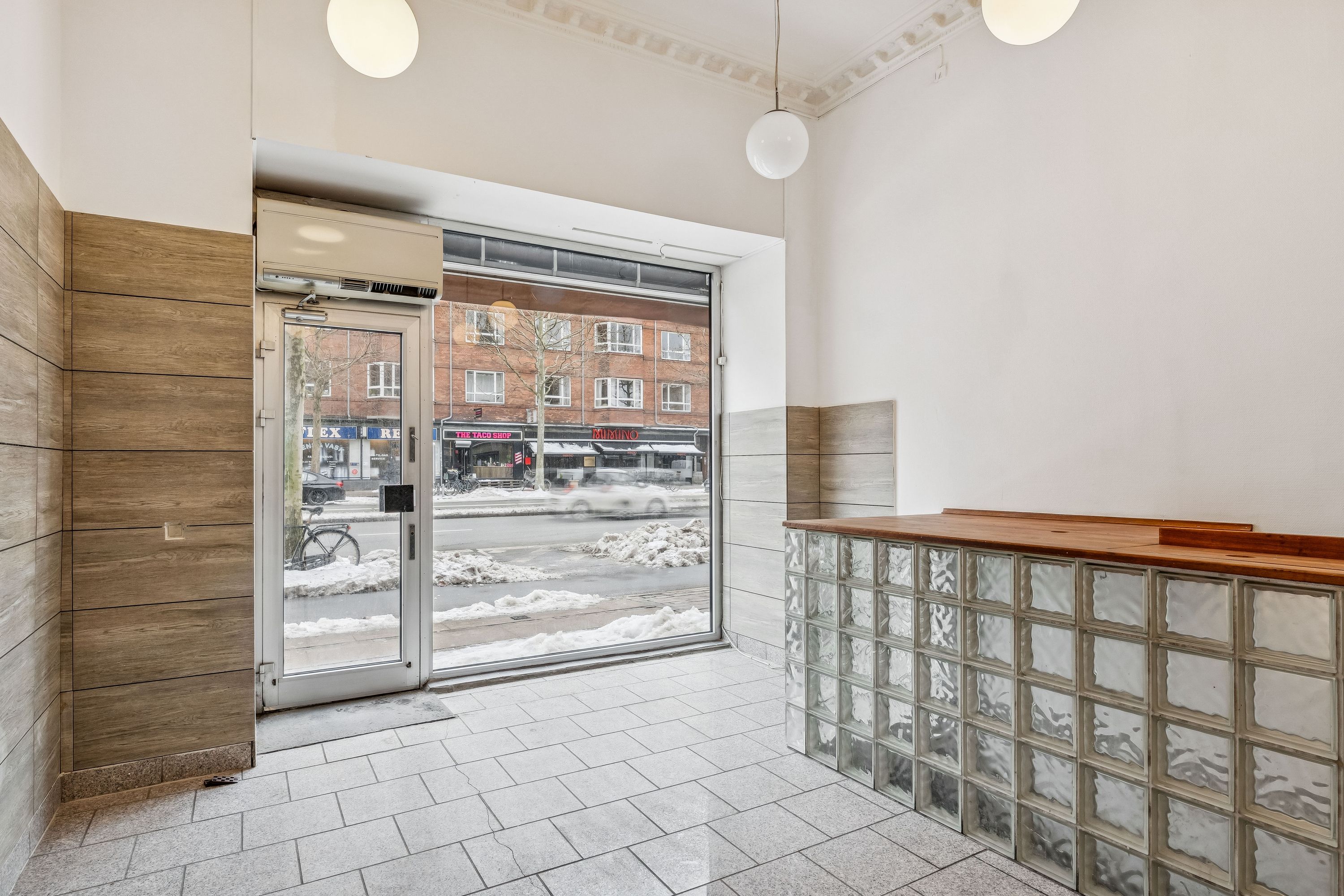 Indvends i en lejlighed med store vinduer og glasvæg, hvor luften strømmer ind. En snelagt vej og flere butikker med skilte synes udenfor. En simpel, men funktionel indretning med træ og glas.