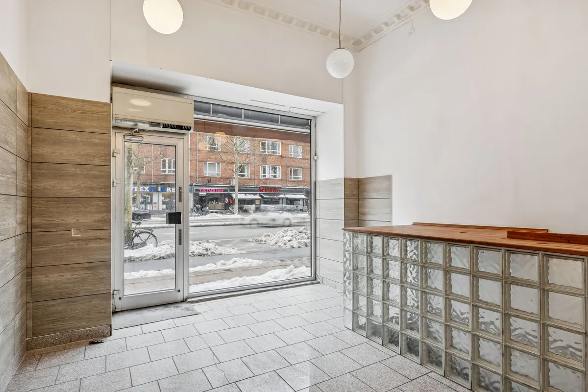 Indvends i en lejlighed med store vinduer og glasvæg, hvor luften strømmer ind. En snelagt vej og flere butikker med skilte synes udenfor. En simpel, men funktionel indretning med træ og glas.