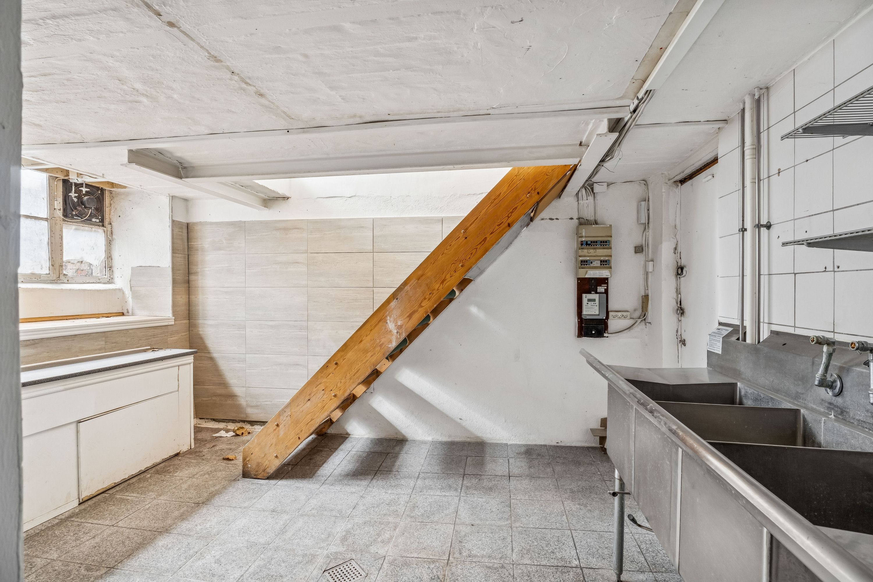 Køkken med skabe, dobbelt vaskebækken og stående trætrappe med ventil, der vender mod højre og har ventilationsskodder i vindue.