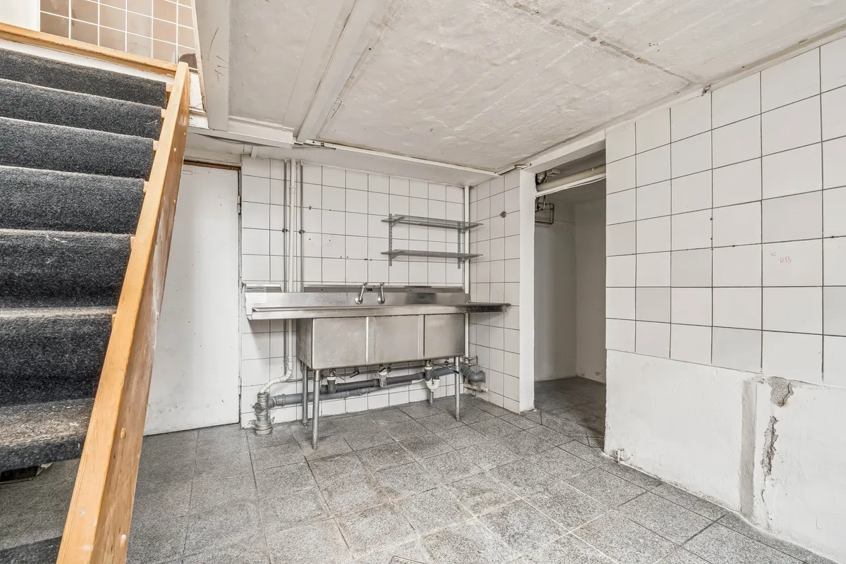 Et køkken med dobbeltvask, metalreol og trappe udenfor, gulve og vægge er snavset og skadet, og der er et hul i højre væg.