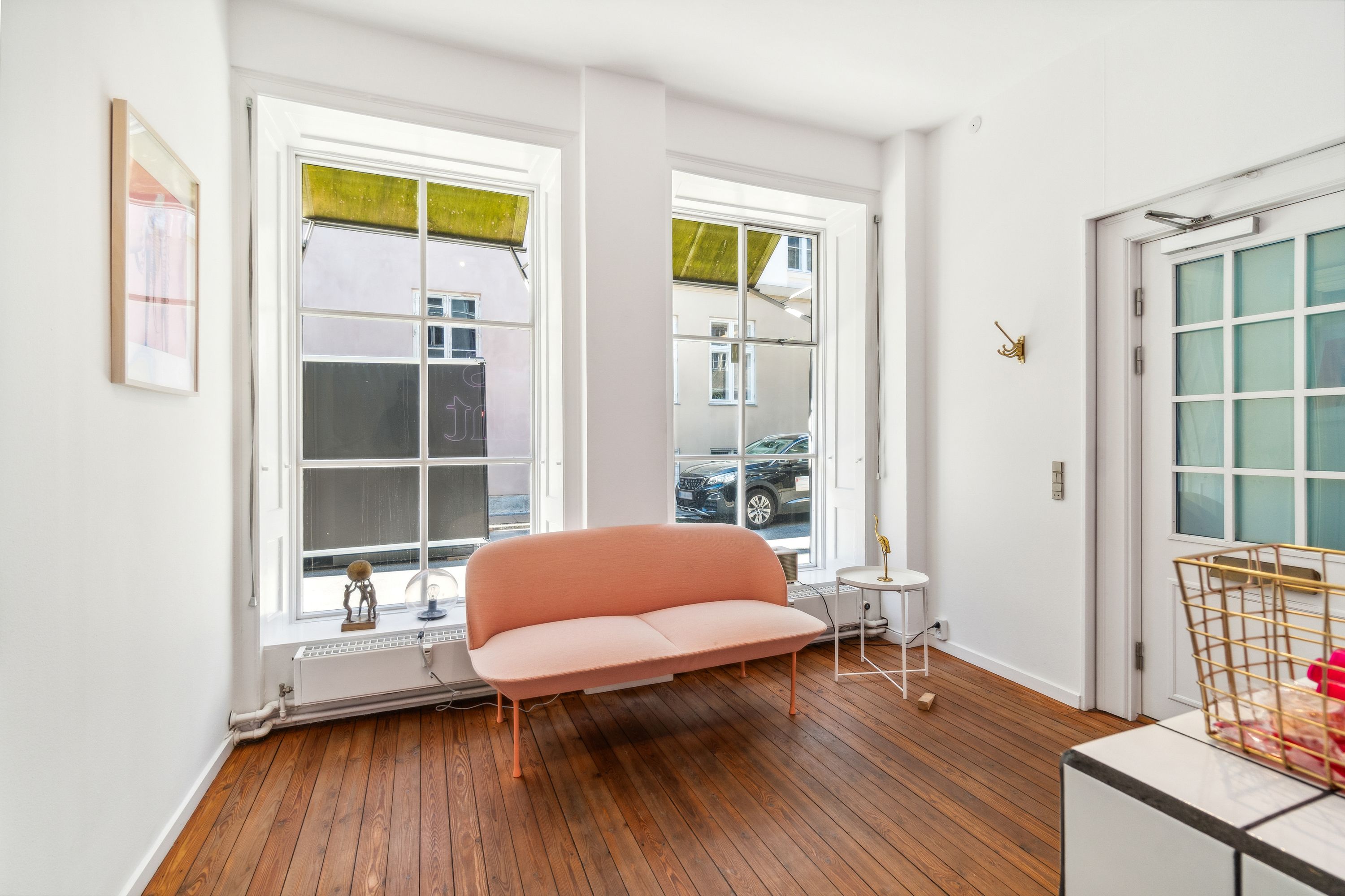 Kontor med lys og moderne interiør, hvor en pink sofa er placeret ved vinduet med udsigt til en bilparkeringsplads og et glasdøre