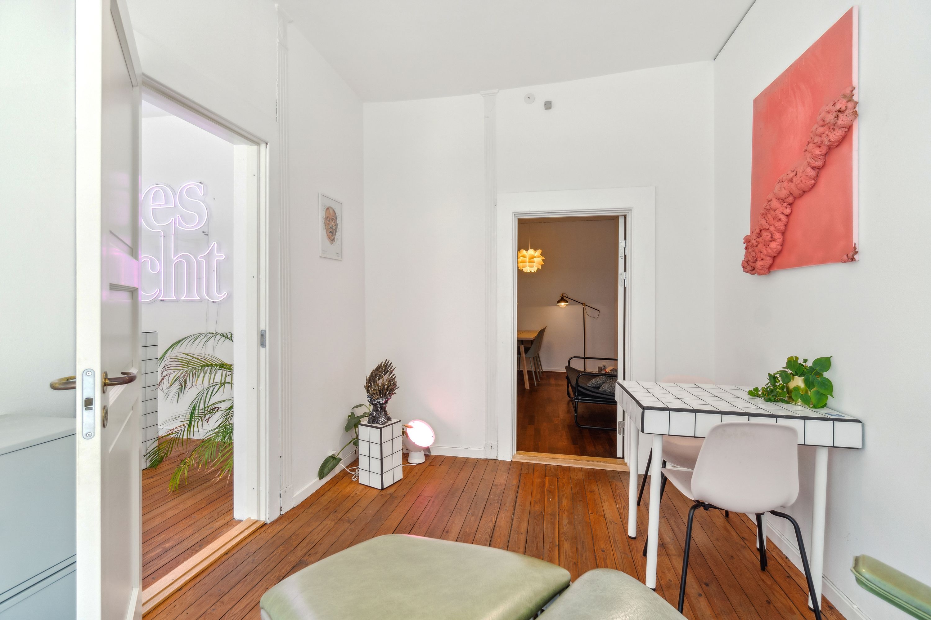 Minimalistisk stue med hvidmalede vægge og massivt trægulv. Vindue med neonlys og lyskrone. Bord med grøn plante og pink maleri. Møbler og tekstiler i hvid og grønne farver