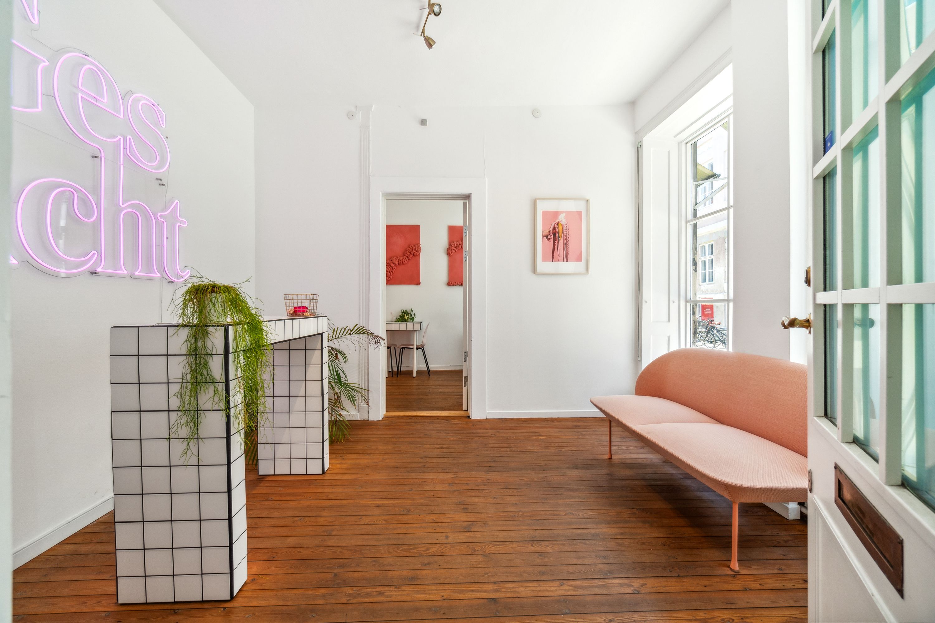 Kontor med rosa sofa og grønne planter. Neon lysskilting med teksten “Rester Chrt” og malerier på væggen. Trægulv og hvide vægge. Vindue med håndtag og udsigt til bygning med cykler.