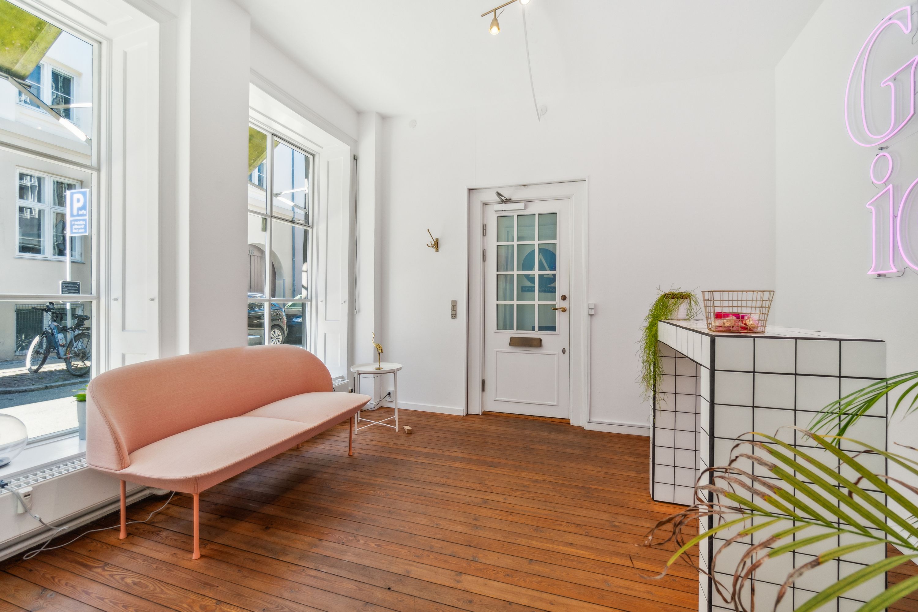 Kontor til leje med en stor lyse stue og moderne møbler. En pink sofa og et hvidt bord med plante i hjørnet. En hvid væg med lysekroner og et pink neonskilt med teksten Grid 10.