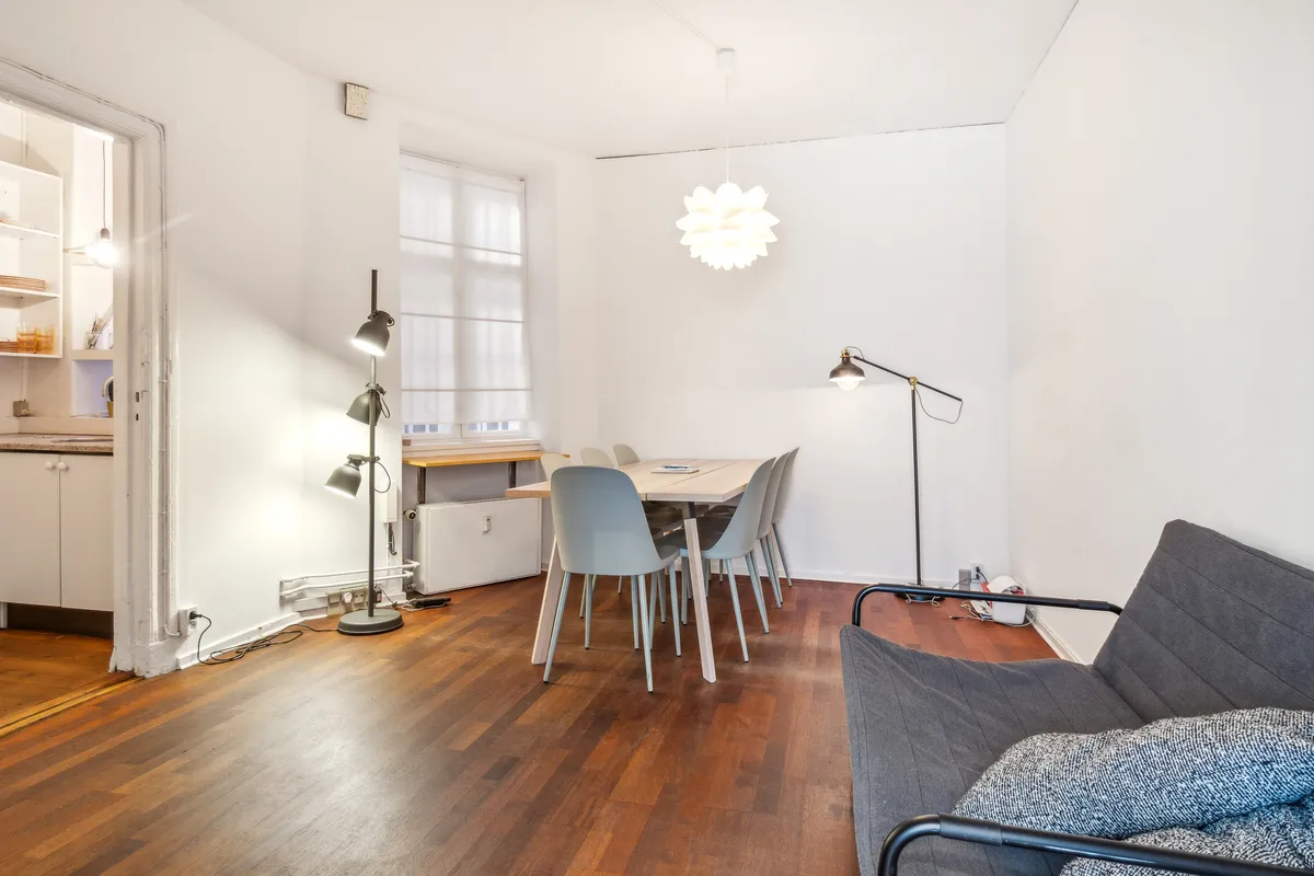 Kontor til leje med åben indgang, bord og stole, og en sofa med pude. Lamper og en sofa er i baggrunden. Væggene er hvide, og gulvet er lavet af træ.