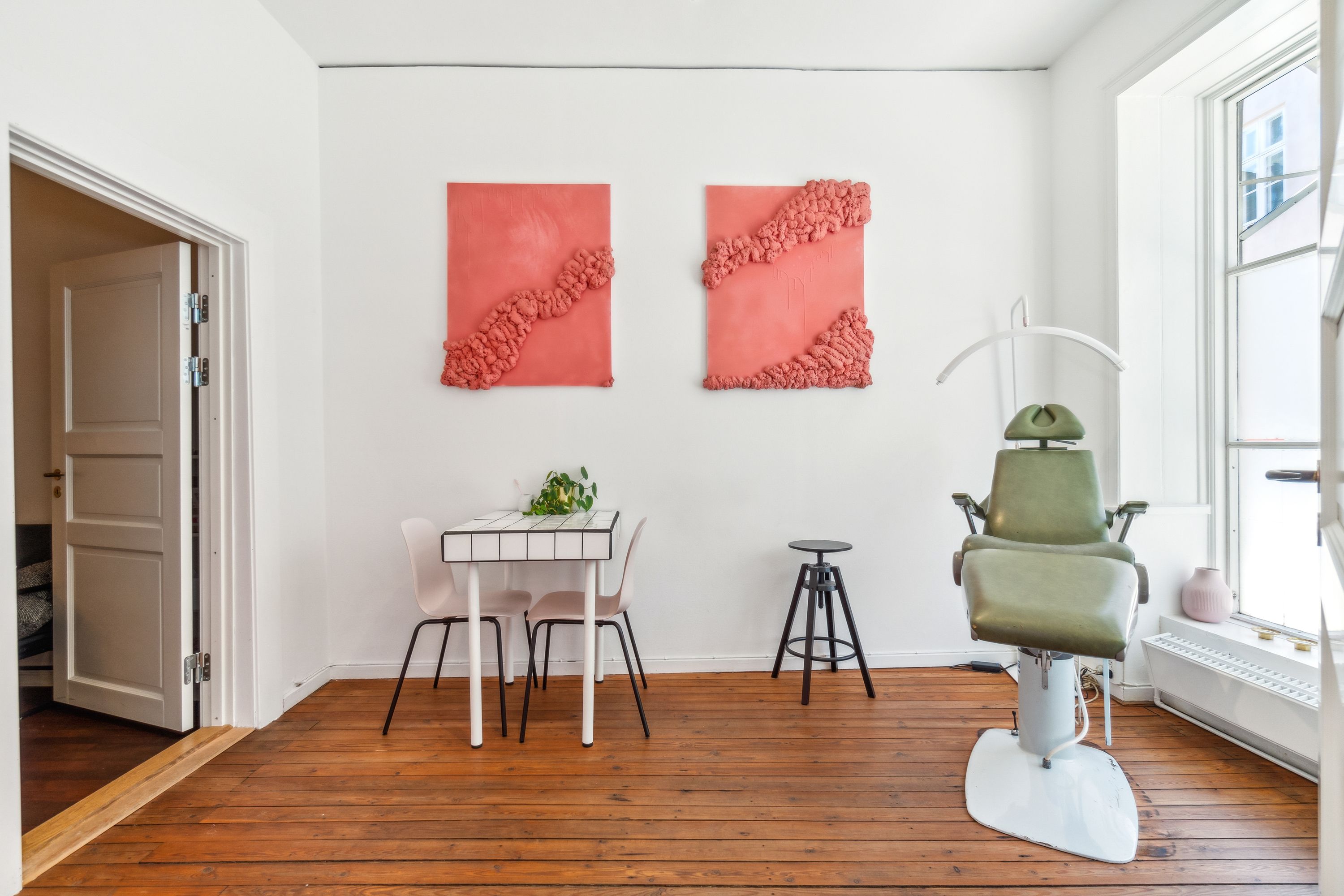 Kontor med højloft og loftslamper, hvid og brun gulvbelægning, lyserøde vægkunstværker, hvidt bord med to stole og en vase, og en grøn lægestol med håndtag