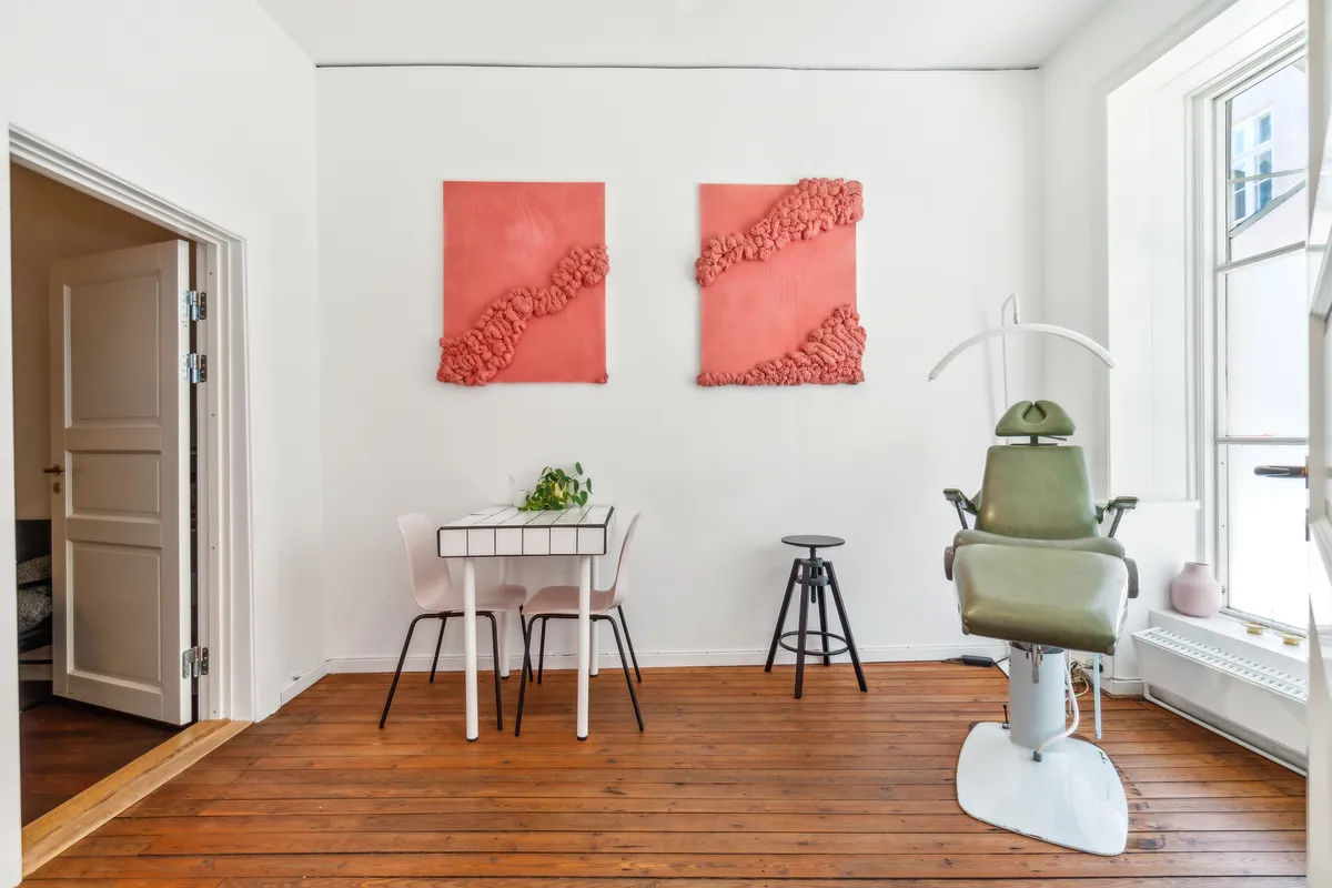 Kontor med højloft og loftslamper, hvid og brun gulvbelægning, lyserøde vægkunstværker, hvidt bord med to stole og en vase, og en grøn lægestol med håndtag