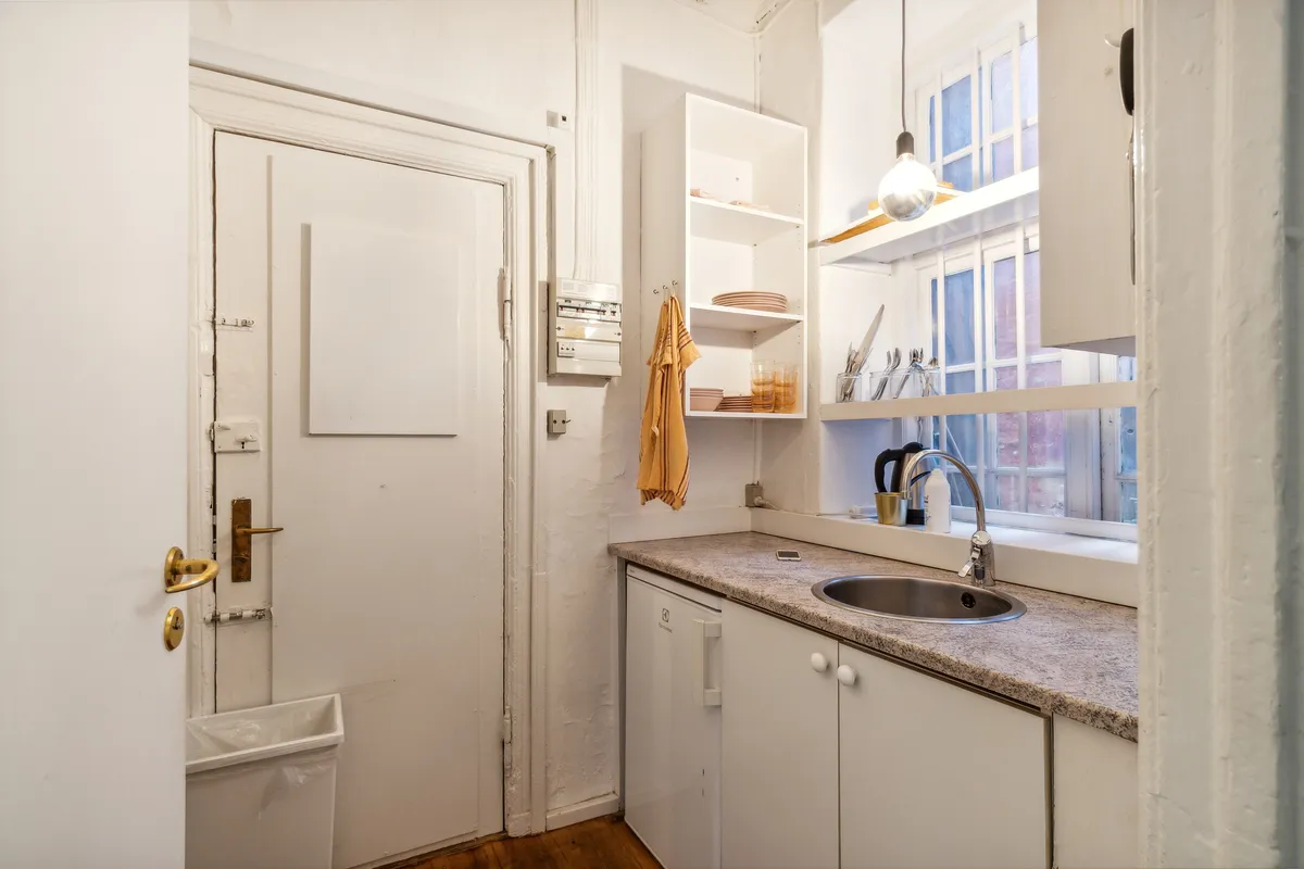 Et rent og minimalistisk køkken med hvide vægge, hvidt loft, granitbordplade, en håndvask med vandhane, et hvidt køleskab og et hængende lys