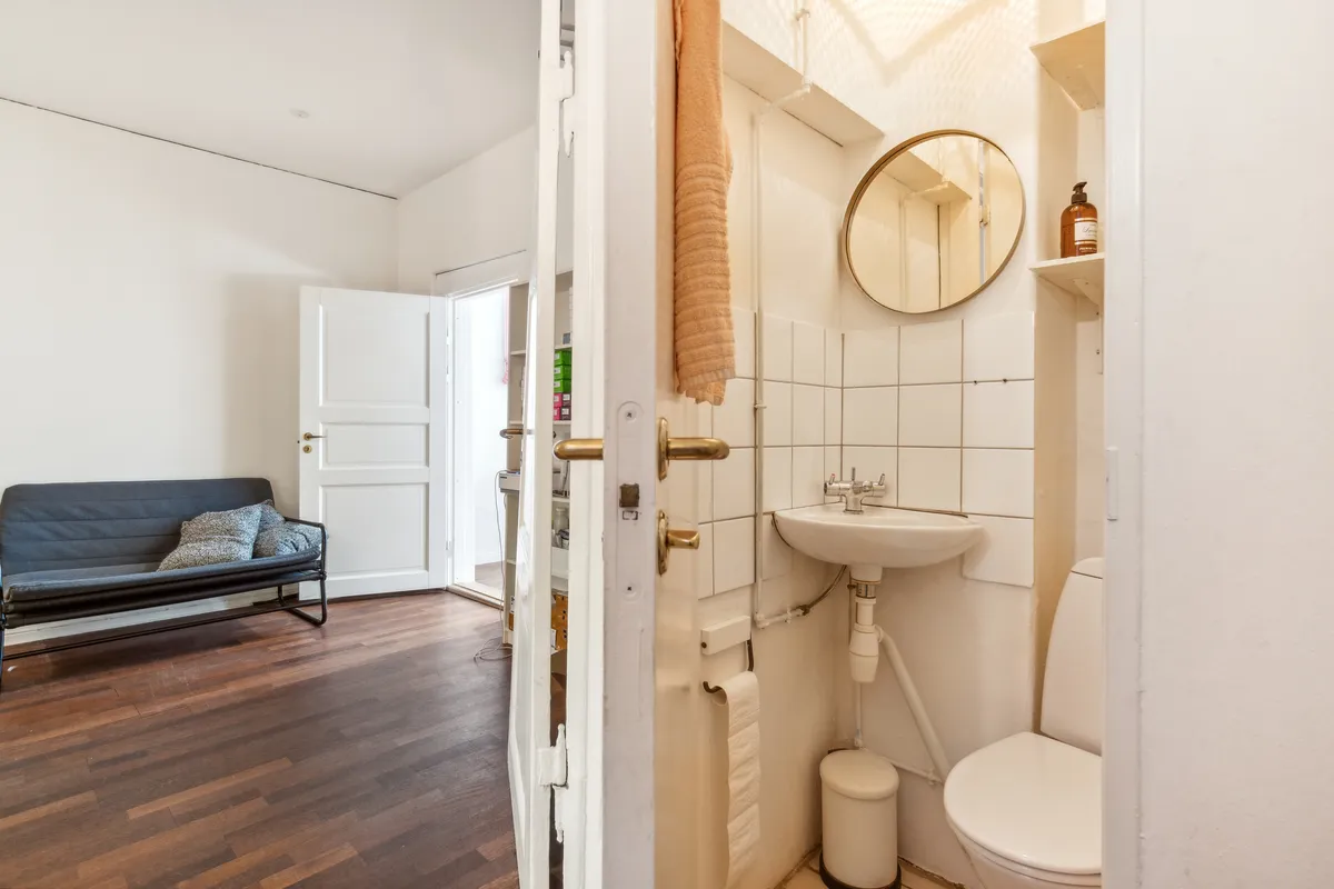 Kontor med en hvid væg og trægulv. Et enkelt sæde med pude placeret på gulvet. Et toilet med en hvid toiletbøsse og en rund spejl.