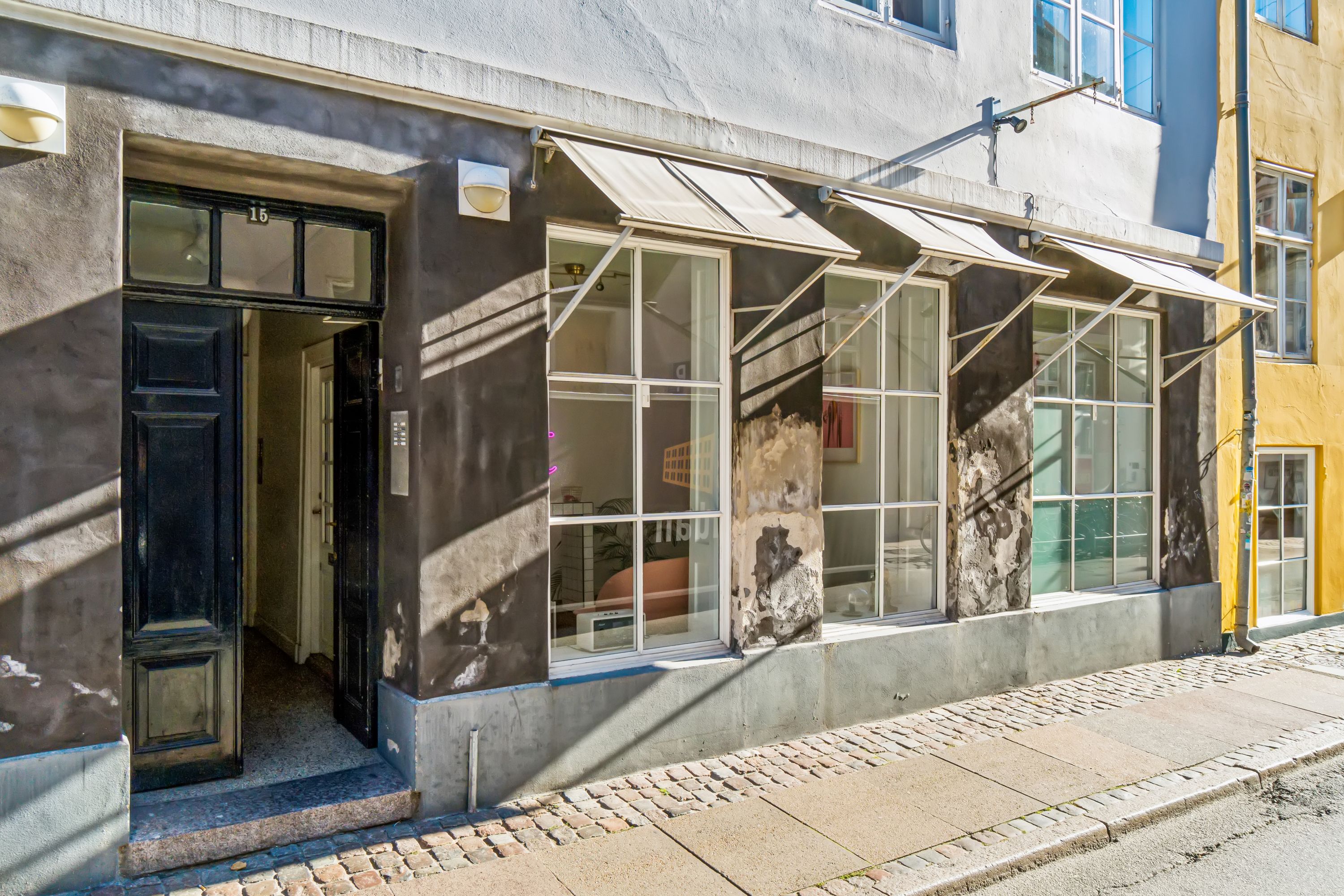 Kontor til leje på Knabrostræde 15 i København. Lejligheden har store ruder, indgangsdør og udvendigt lyskab. En af ruderne er åben og viser et rum med møbler og udstyr.