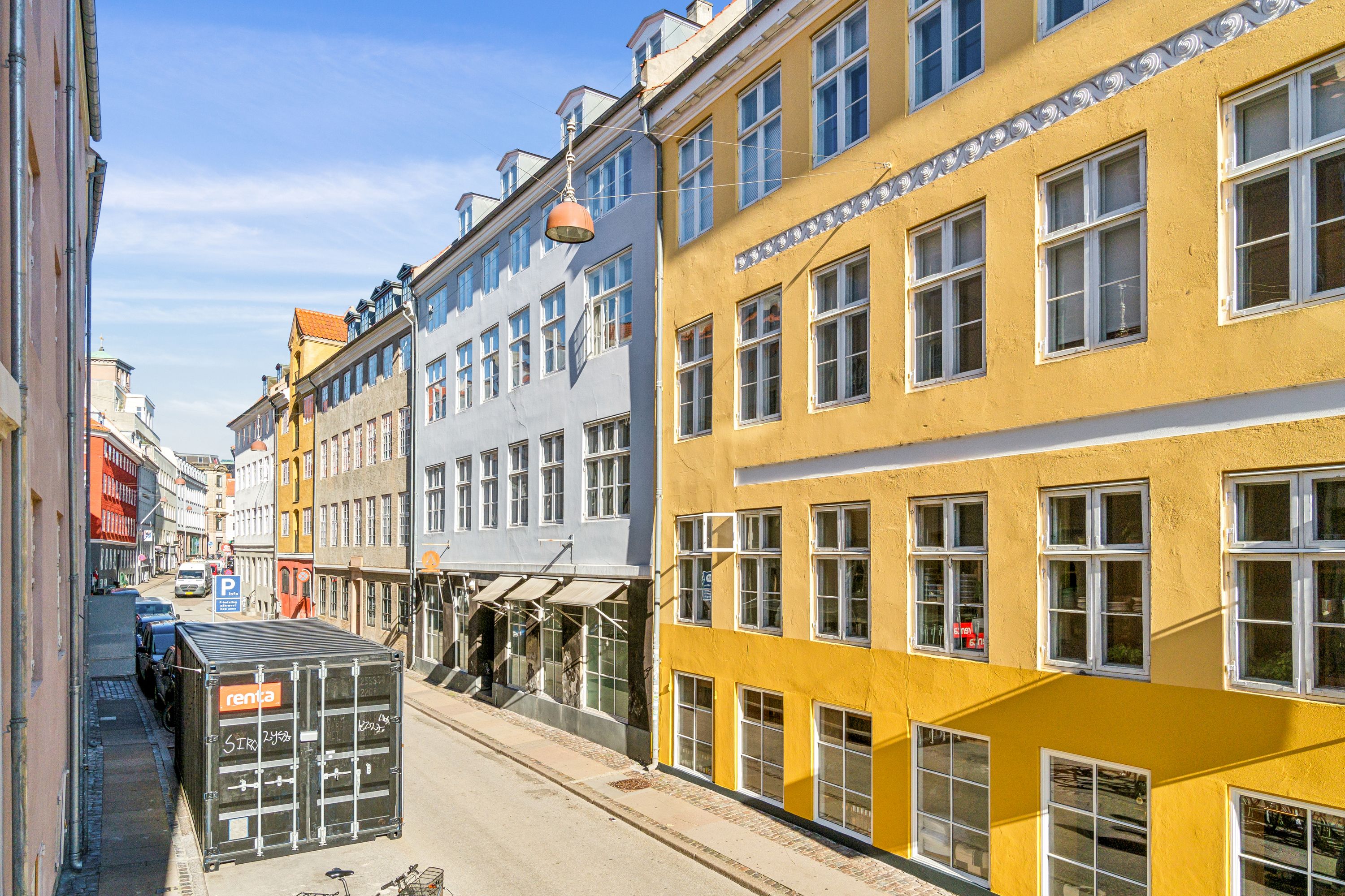 Lejlighed til leje i Knabrostræde med smukt gult facade og mange vindue i stueetagen. Parkerede biler og en container foran lejligheden