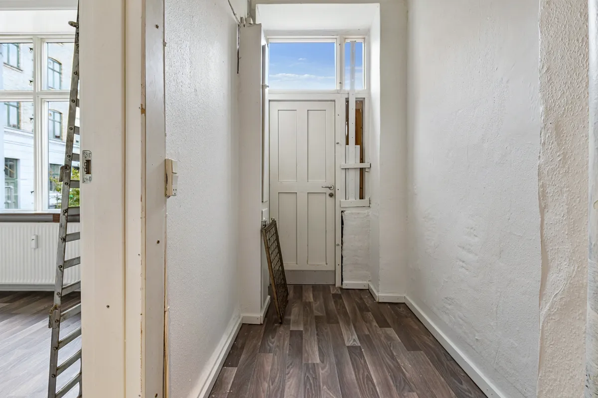 Foto af en smal korridor med hvide vægge, møbleret med et hvidt dørblad og et lille vindue. Gulvet er lavet af træ og der er en stålstige i baggrunden.