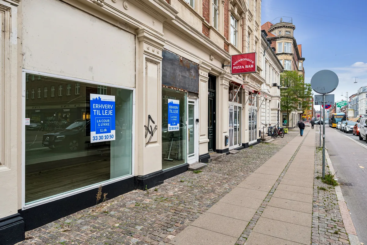 Butik til leje på Amagerbrogade med facadevinduer og skilt, Pizzabar og vagtelskere i baggrunden, personer og biler på gaden