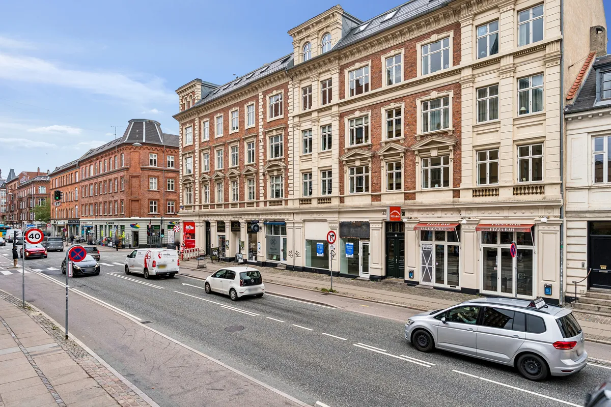 Menighedsgården på Amagerbrogade i København S er opdelt i flere lejligheder og butikker. Foran er der en bilparkeringsplads og flere biler kører langs gaden