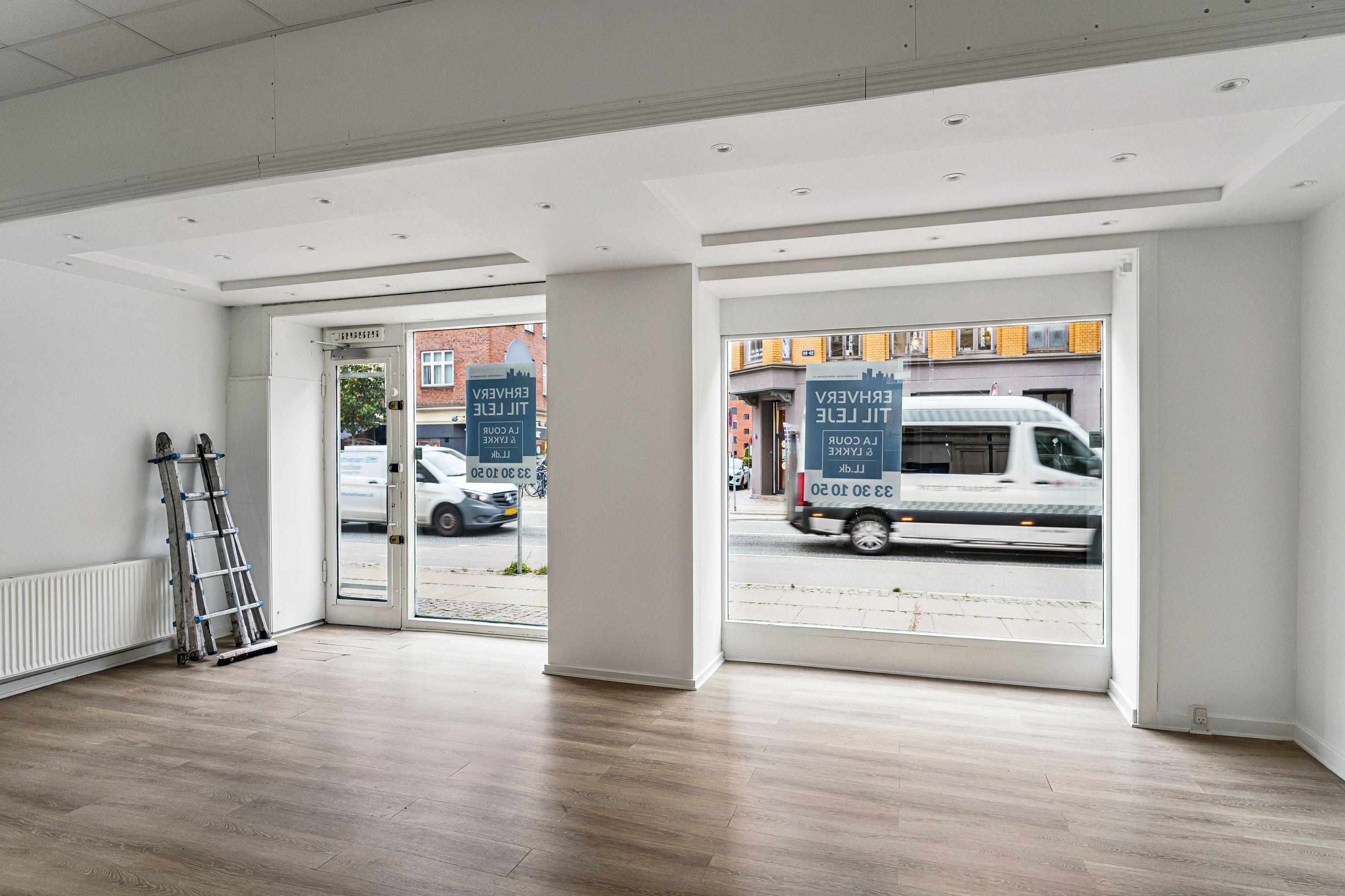 Lukket butik til leje med to store glasvinduer og lyse lofter. Stående skilt med "FOR RENT" og "FOR LEJE" i gul skrift på hvide skilte. Biler kører forbi på Amagerbrogade.