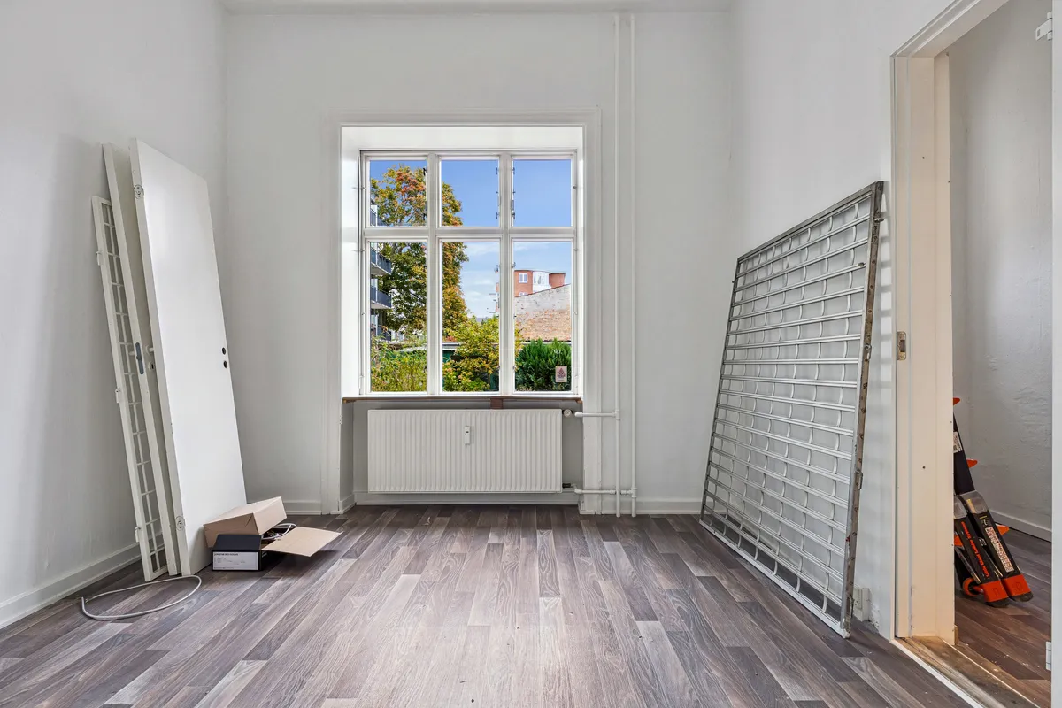 Lukket salgslokale med åbent vindue og lyse hvide vægge, trægulv og radiatorer samt lukket gardin og kasse med skrift i en tæt stue