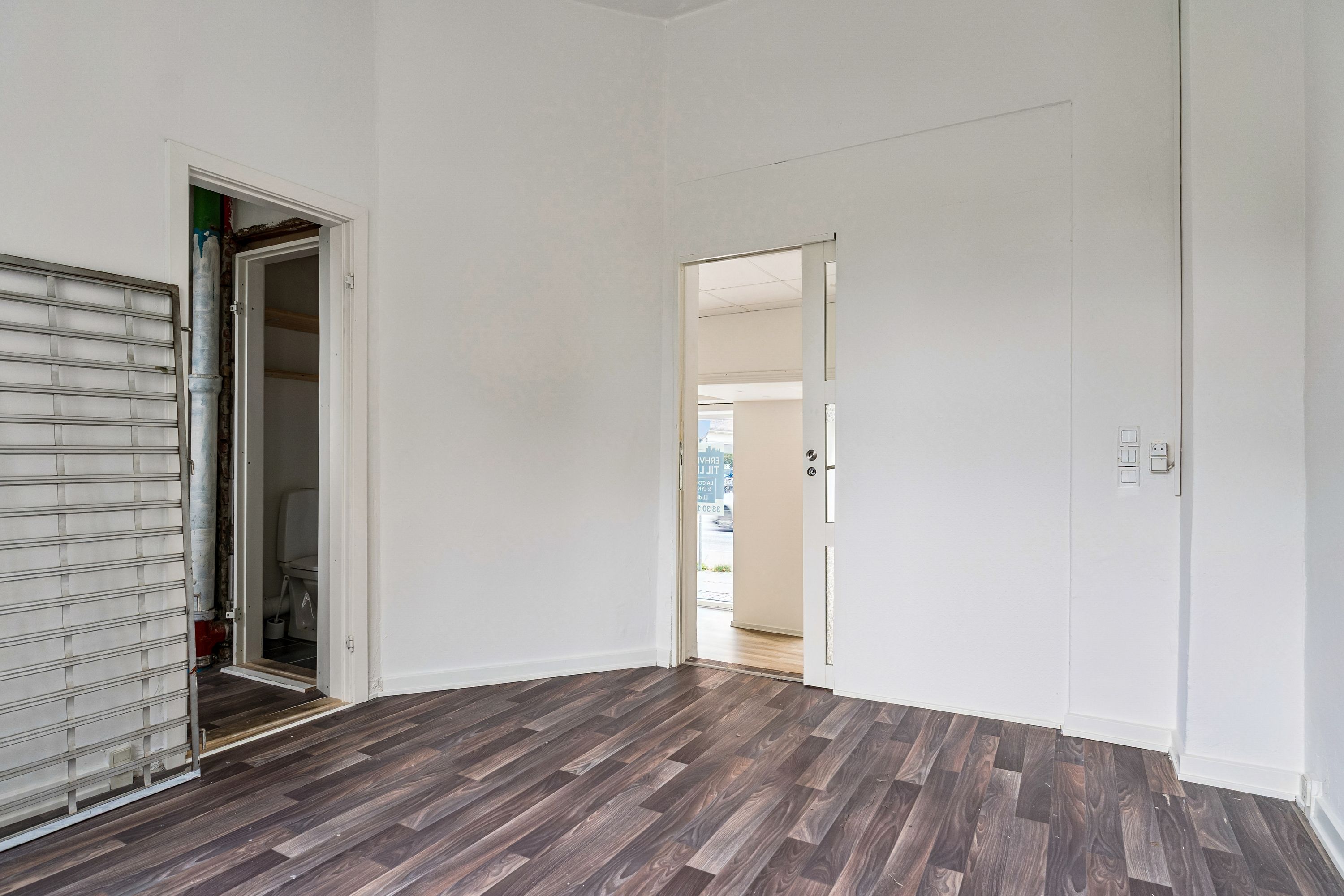 Et tomt lokale med åbne døre, hvor der er højloft og trægulv, samt et lukket badeværelse med toilet og opvask. Der er udsigt ud i gadedannelsen og der er en opbevaringsrum i siden