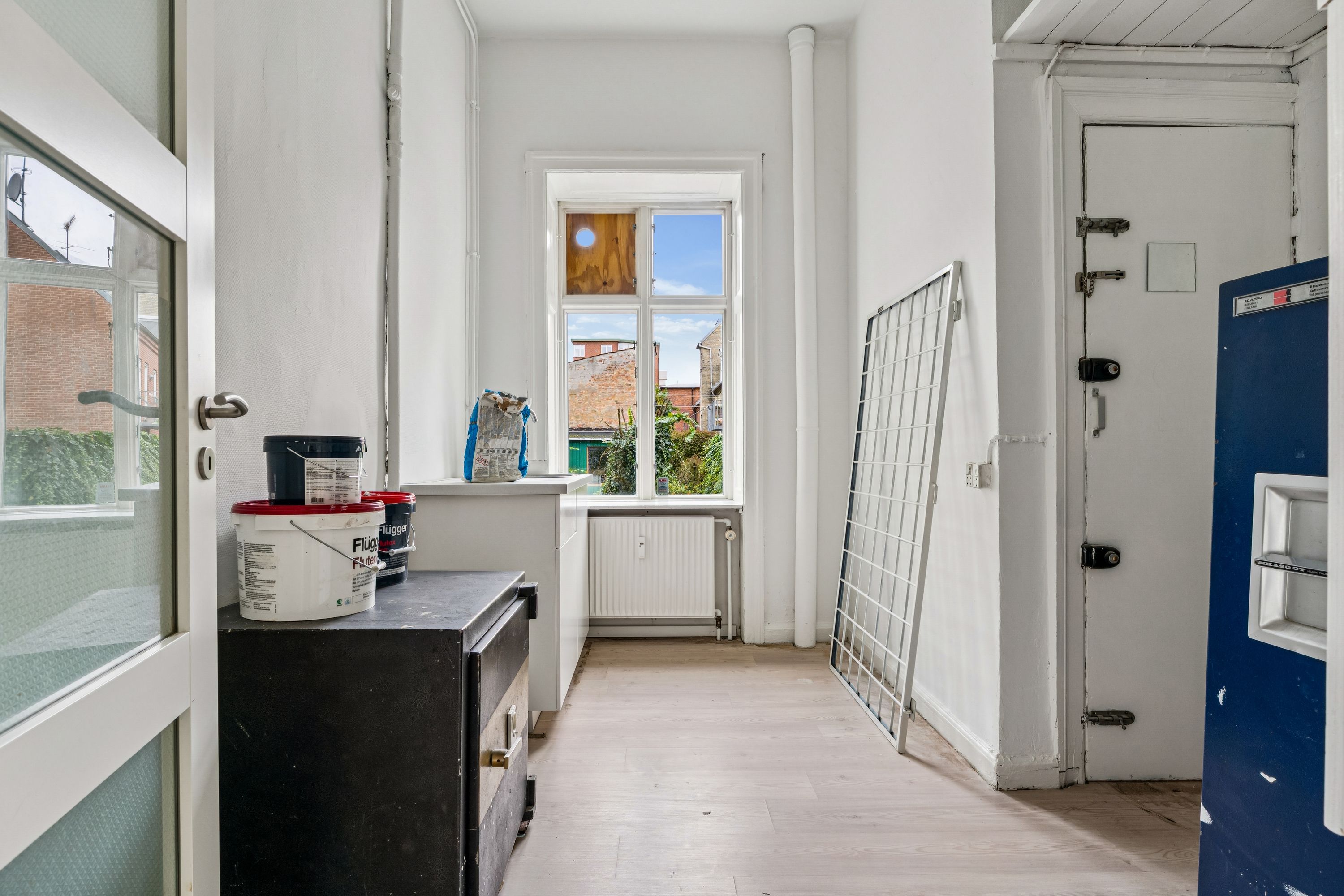 En tom, hvidmalet butik med et salgslokale, trægulv og et lagerrum med flere malingsspand samt et radiatorsystem i baggrunden