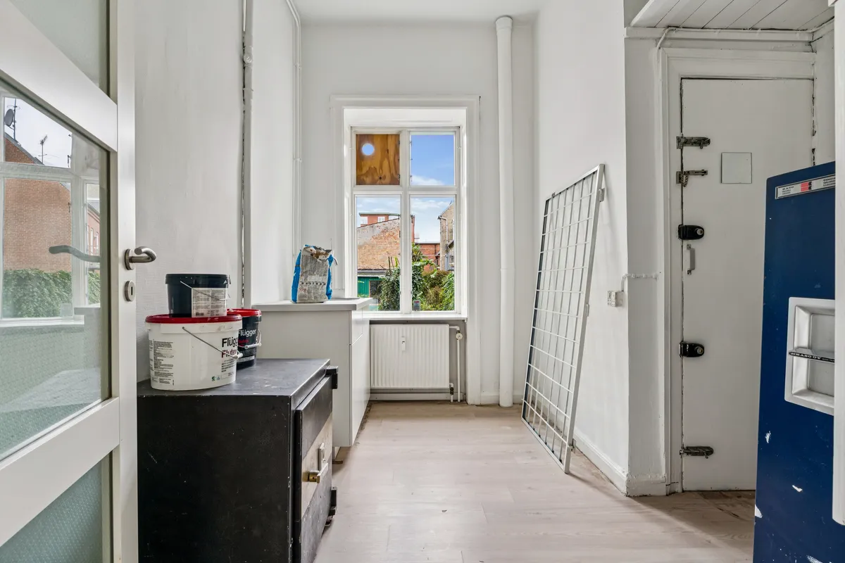 En tom, hvidmalet butik med et salgslokale, trægulv og et lagerrum med flere malingsspand samt et radiatorsystem i baggrunden