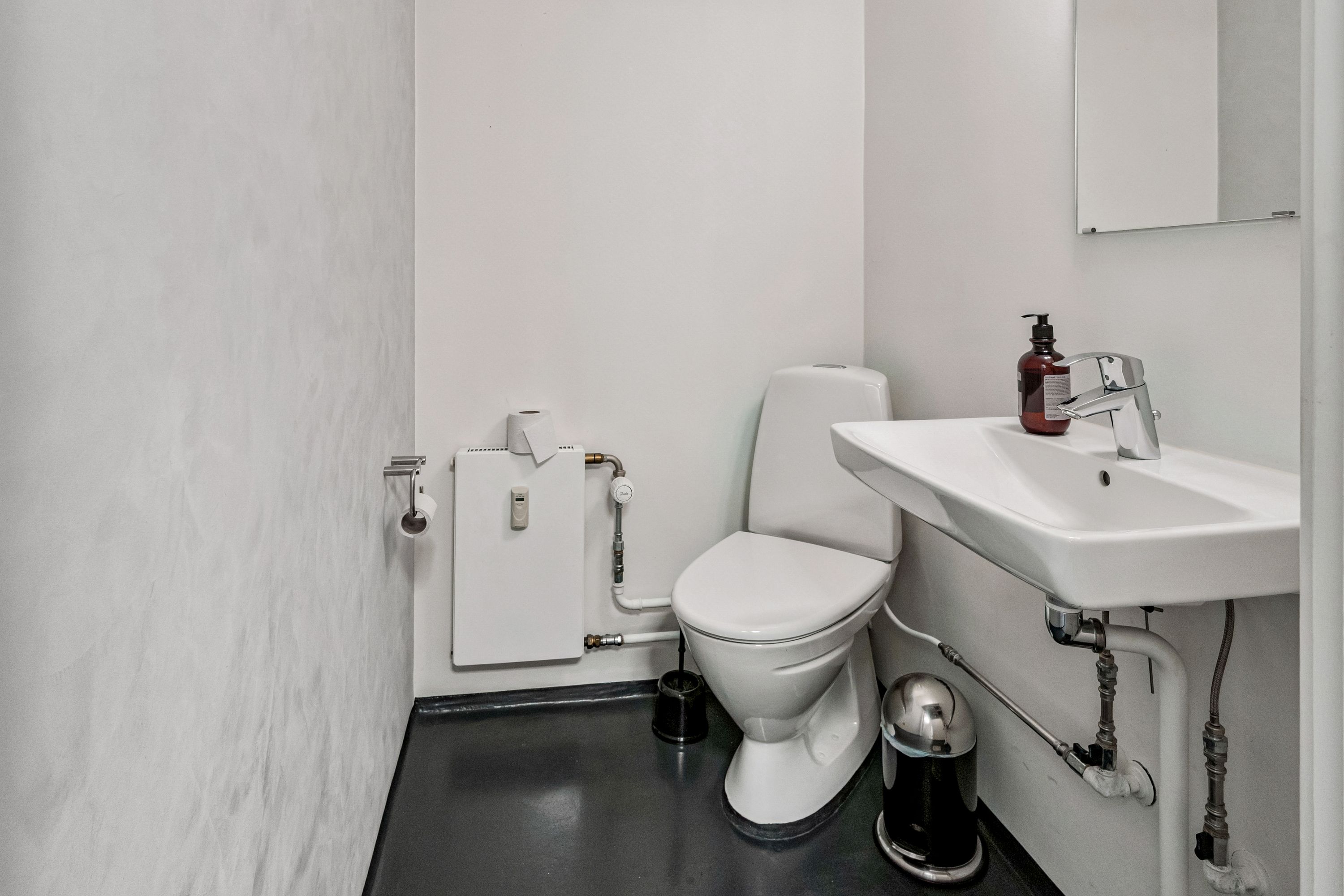 Kontor med toilet og hvidt vask i toiletrummet. Væggene er hvide, og der er en håndklædeholder på væggen. Toiletpapirruller er ophængt på væggen.