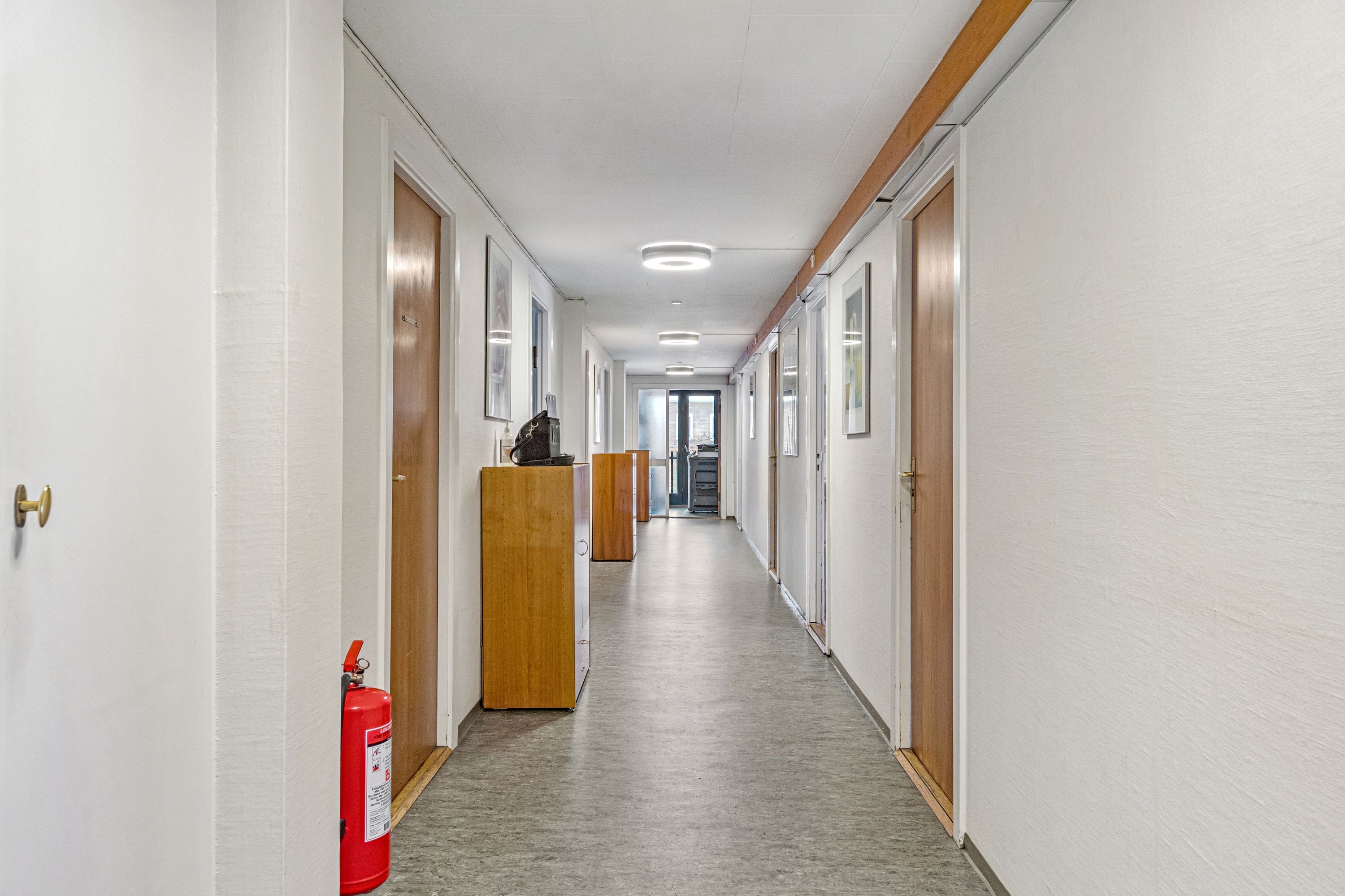 Kontorgang med højt til loftet, lysekroner, trædøre, kabinetter og rødt brandslukker. På højre side er der en dør, og på venstre side er der en dør med guldfarvet lås.