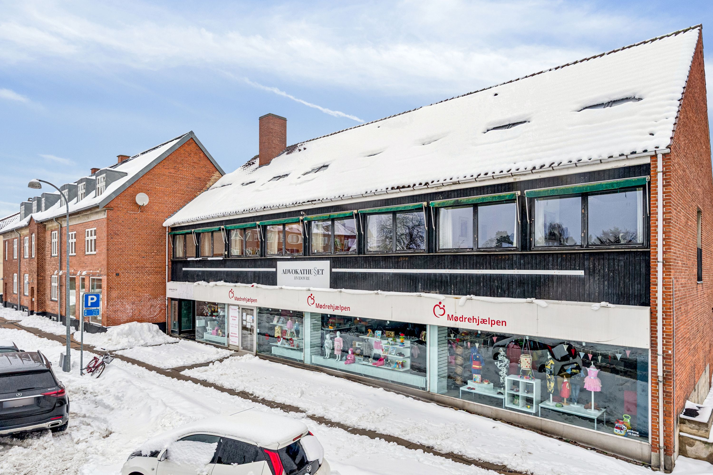 Hvidovre torv 9 med kontorer og butikker. Kontorlokalen er dækket af sne. Biler og en cykel er parkeret. Vinduerne har reklamer.