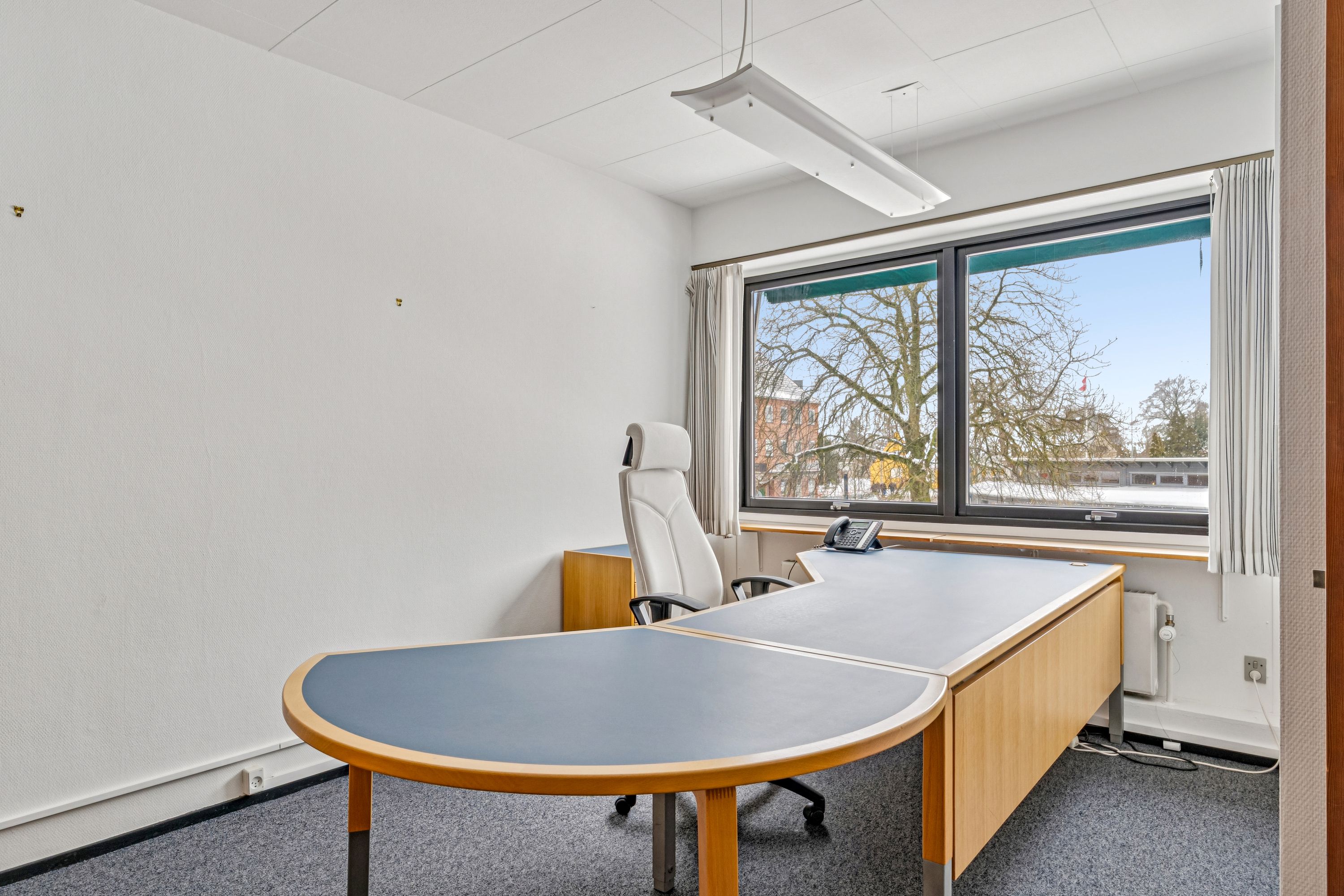 Kontor med hvidt loft, trægulv, U-formet skrivebord, hvid stol, telefon, slank lampe og vinduer med udsigt over træer og bygninger