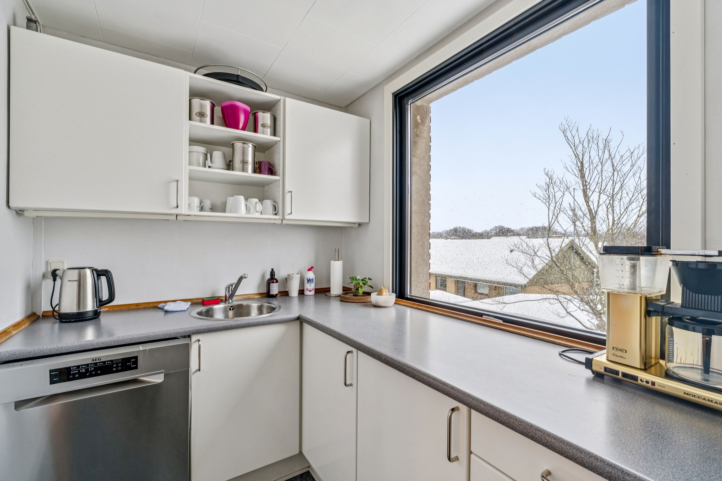 Kontorlokaler med køkken med hvidt køkkeninventar og udsigt til gård med træer og hus. Køkken med grydefad, gryderet, kaffemaskine, strygejern, køkkenhåndklæder og plante.