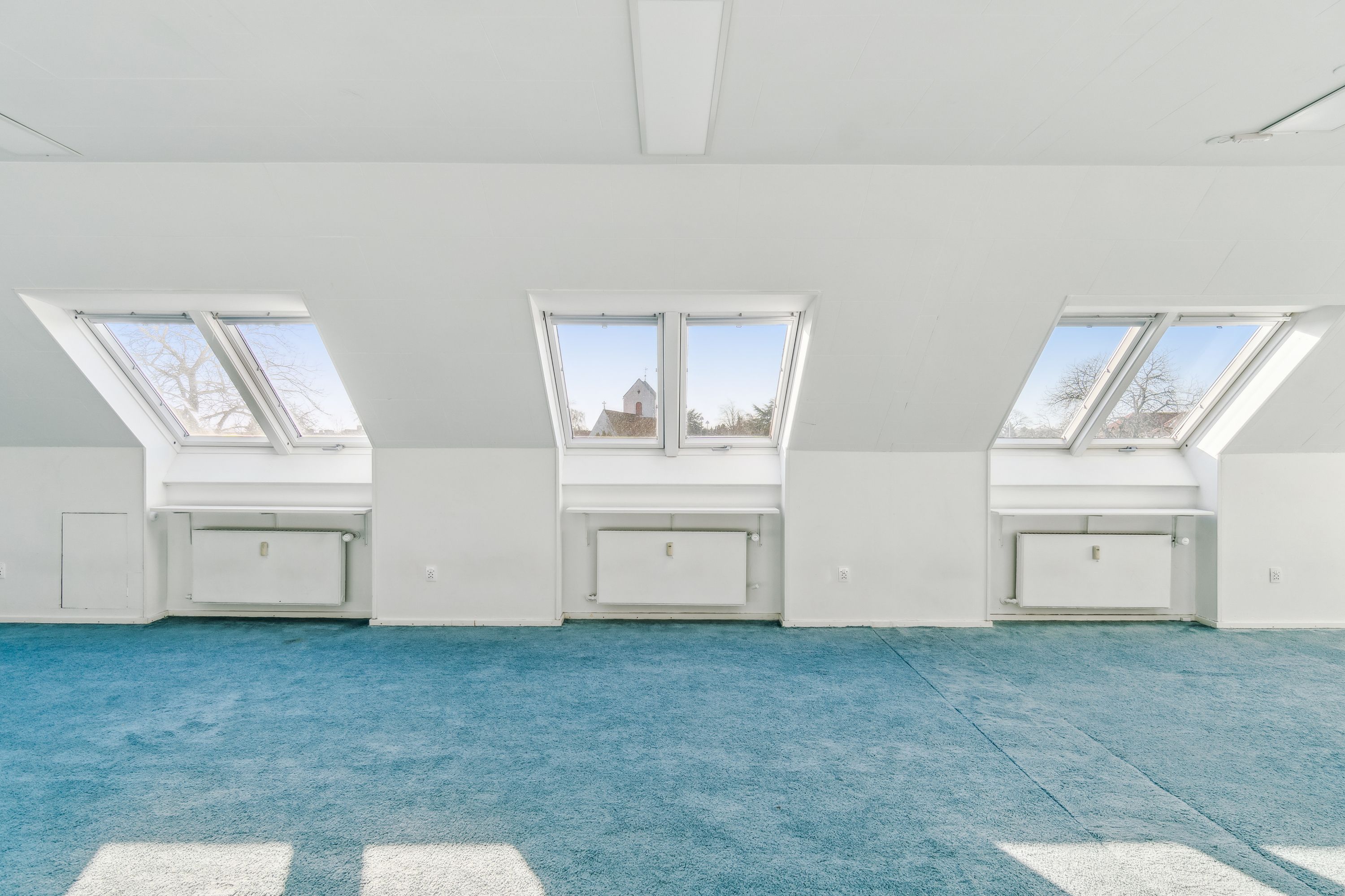 En stor, lyse og ren kontorrum med blå gulvtæppe og tre loftsvinduer med udsigt over Hvidovre Torv. Under vinduerne er der hvide radiatorer og i hjørnerne er der lyse, hvide vægge.