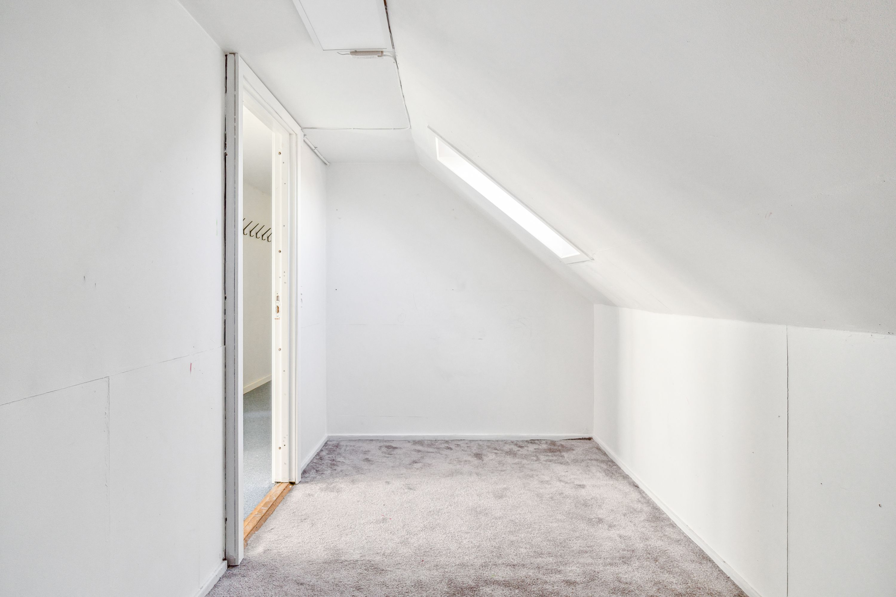 Ledig kontorrum med hvide vægge og loft, gulvtæppe og lyse lyskappe på loftet, der er en åben dør til et rum med en hængeskab