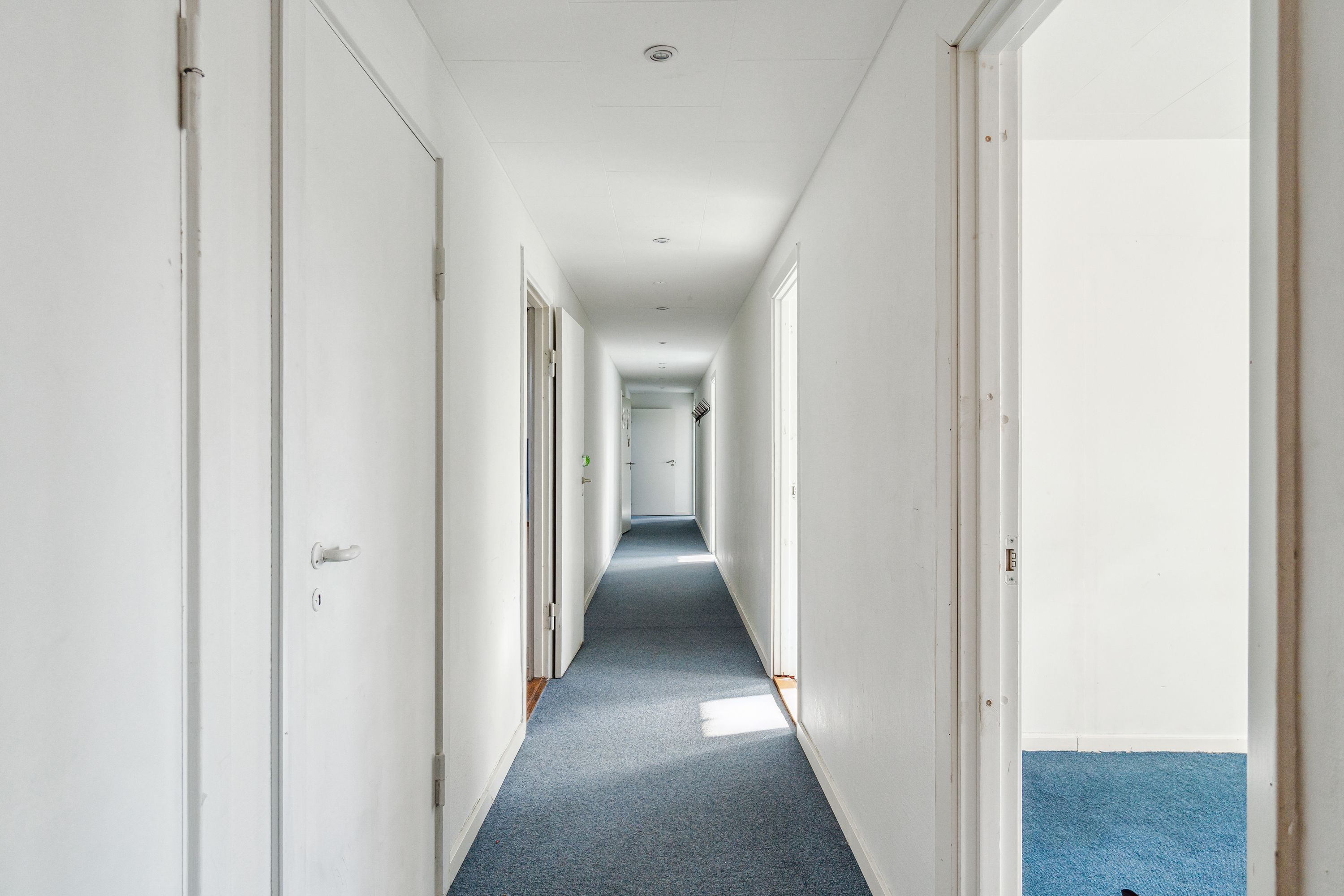Længdevis er der et langt korridor med hvide vægge og blå gulvbelægning. Der er åbne døre og flere vinduer, der giver udsigt til andre korridorer. Der er ingen møbler eller folk på billedet.