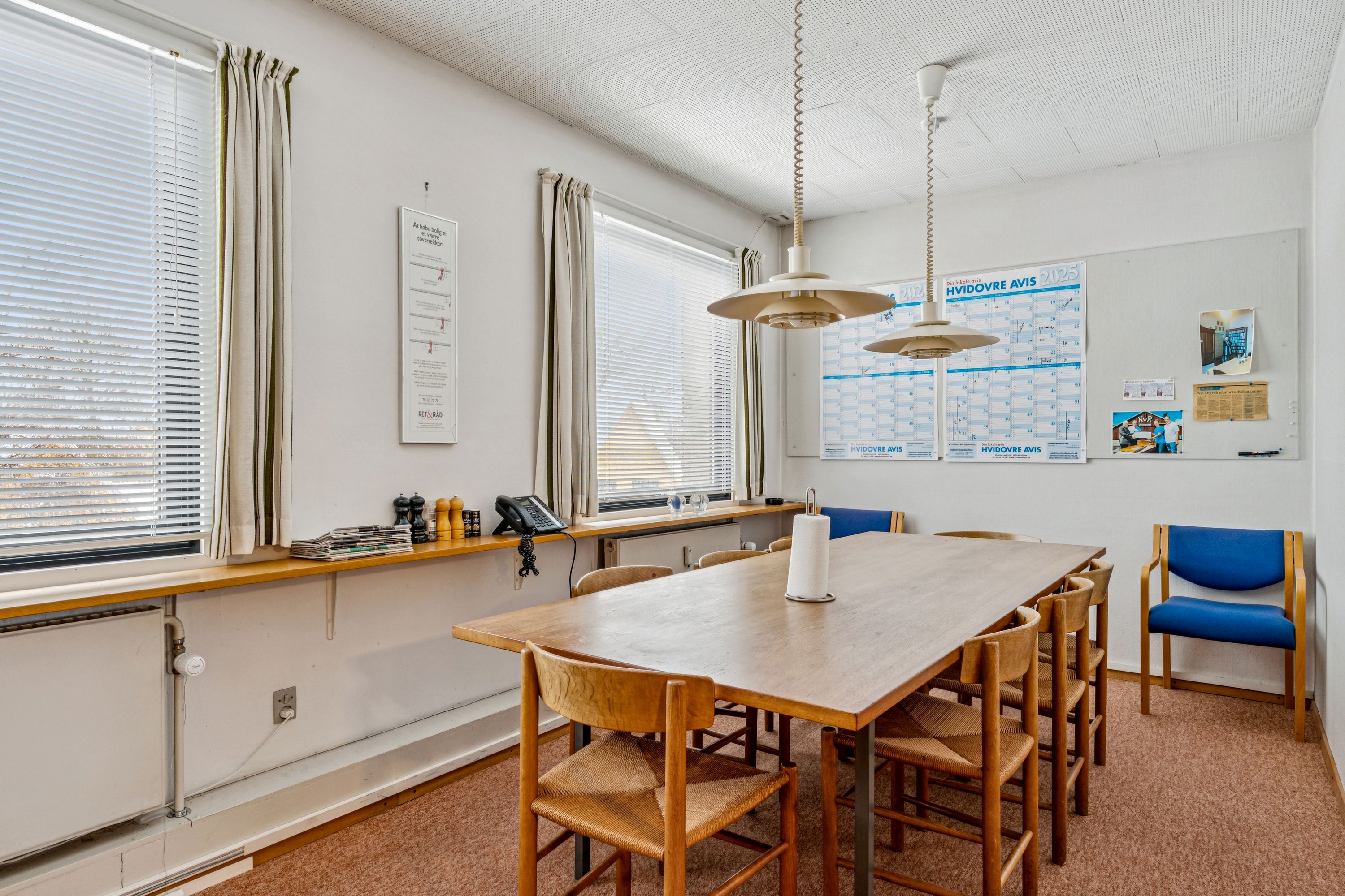 Kontorrum med lyse vinduer og lysekroner, møbler og kalender og plakater på væggen, hængende hvidt papir med billeder og tekst, træbord og stålstole
