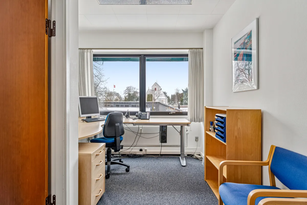 Kontor med skrivebord, stol, computer, telefon og hyggeligt udsigtsvindue. Møblerne er i naturlige farver og rummet er lyst.