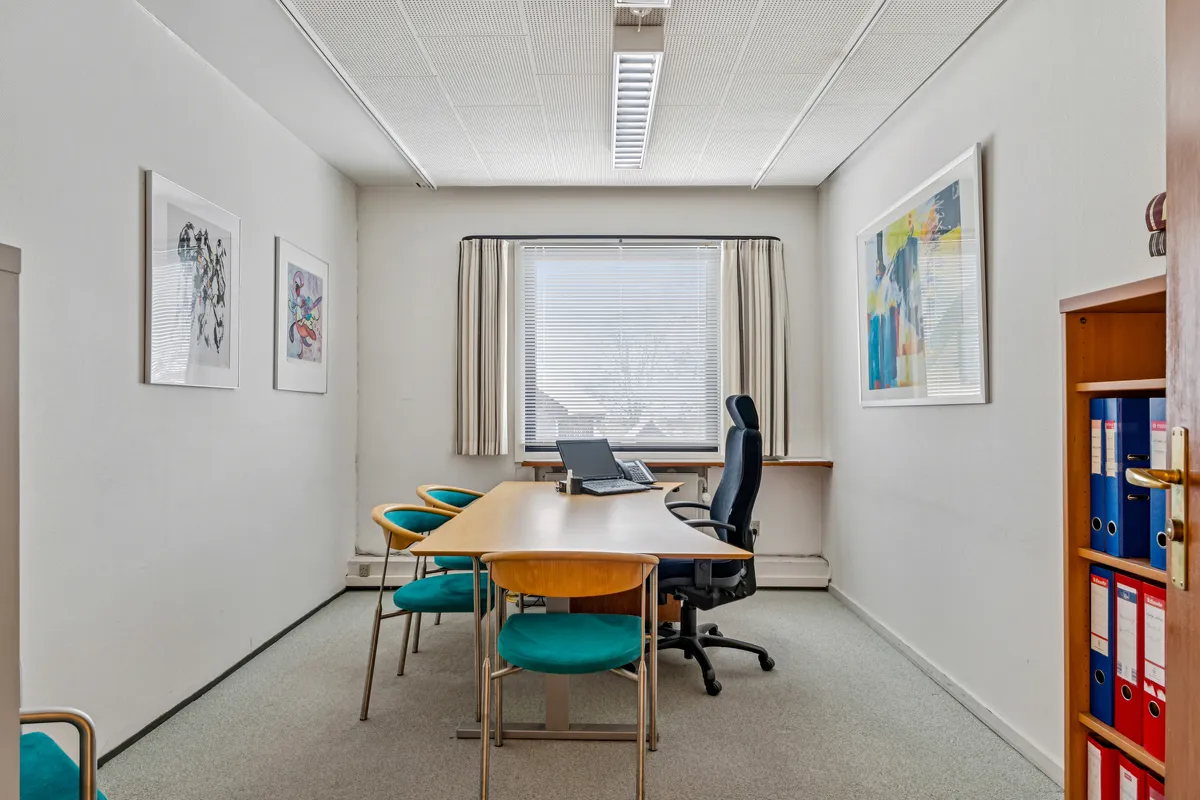 Kontorrum med skrivebord, kontorstole, fire stole, og et lille hyldekabinet med mapper i en hjørne.