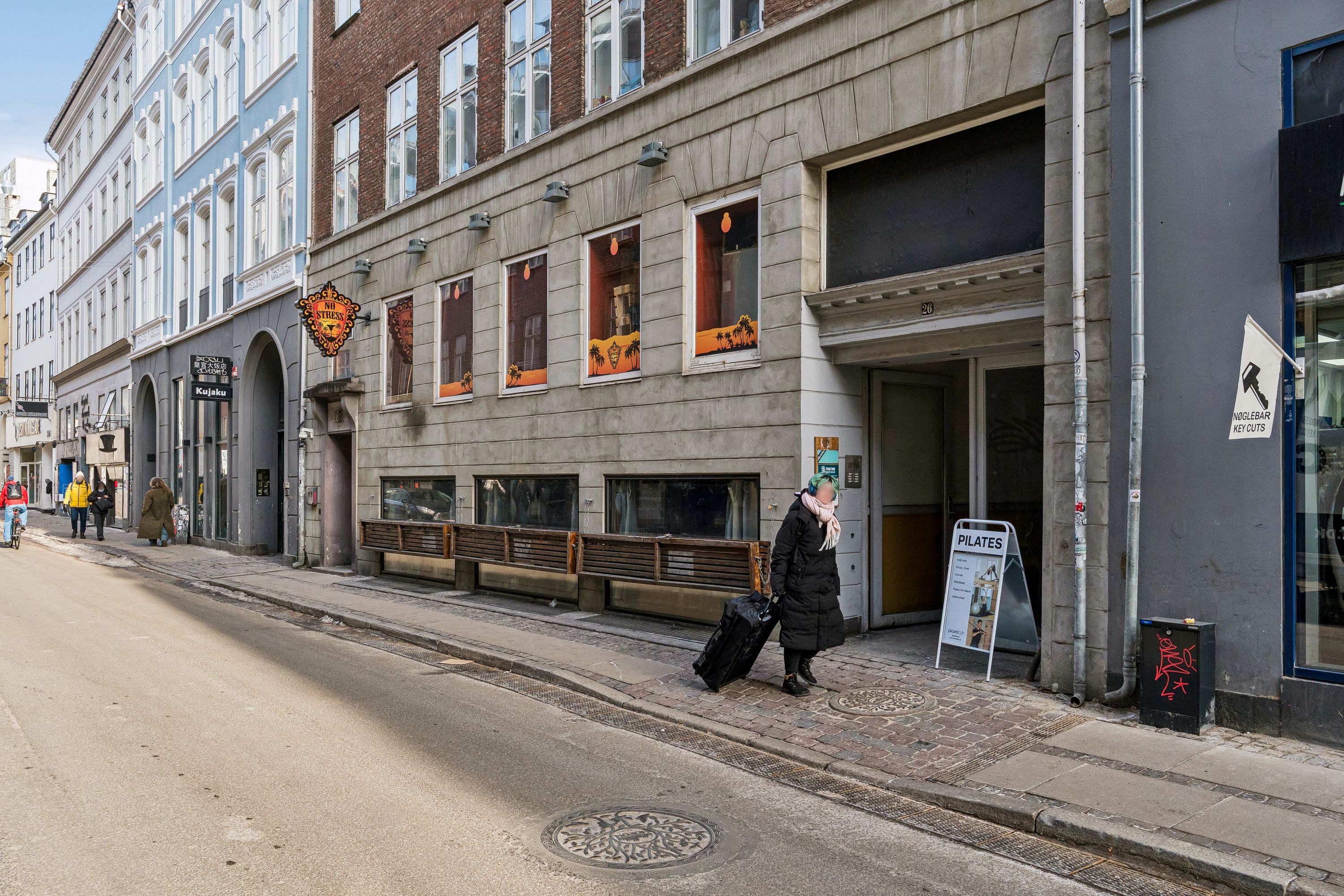 En kvinde går med kuffert og håndtaske foran Nørregade 26. Kontoret har moderne møbler og lyse vinduer med udsigt til gaden.