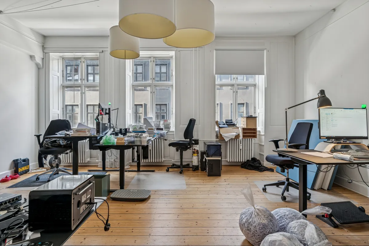 Kontor med lyse vinduer og lysegule loftslamper, computere og skriveborde, hvor der er plaget papposer og emballage.