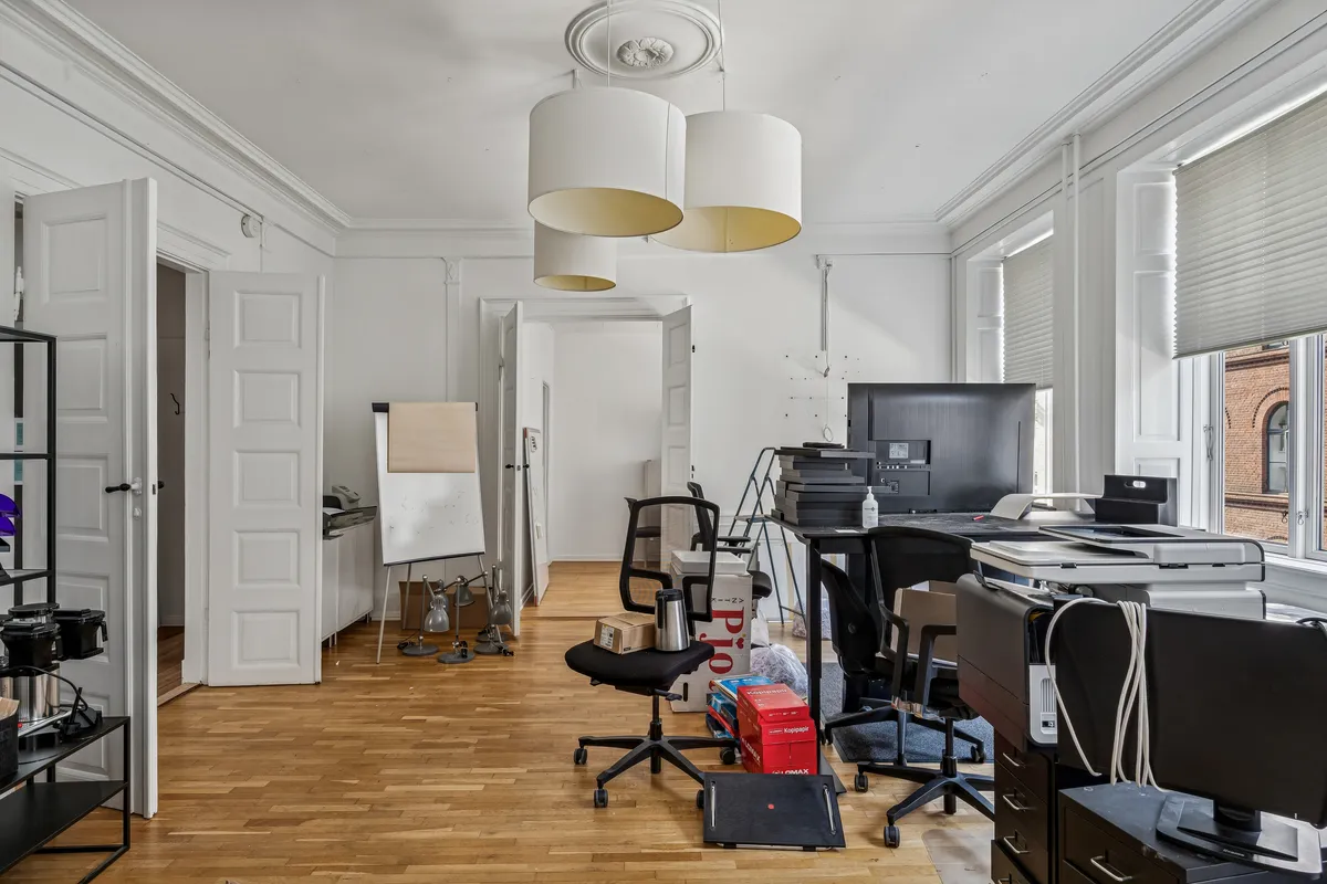 Kontor med højloft, hvor der står møbler og elektronisk udstyr samt en stol med en kuffert. Der er 3 lysekroner i loftet og et vindue med gardiner