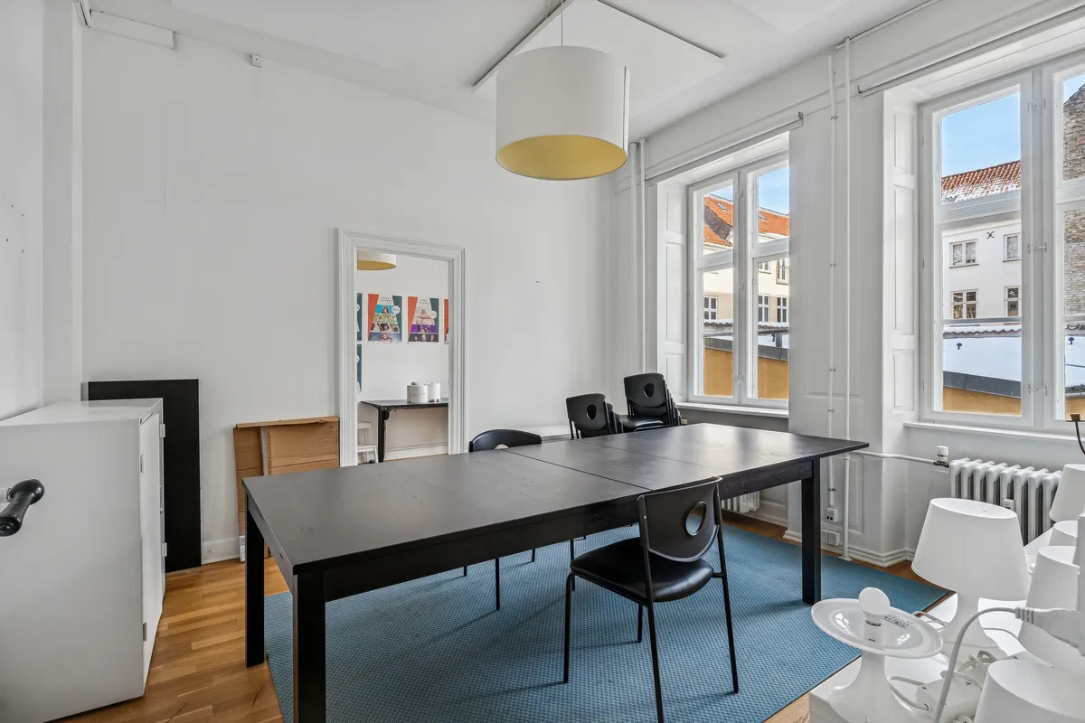 Lyst kontor med højloft og lyse vinduer, møbler, skriveborde, stole og lampe. Lejligheden har lyse vægge og trægulv, vinduer med udsigt til Nørregade og bygninger.