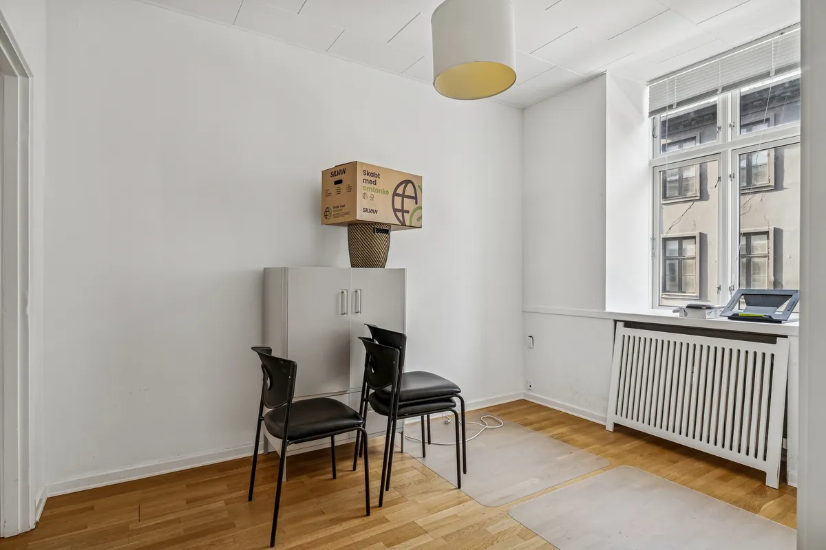 Lejemål med lyse kontorer og højloft. To stole og et højloftsskab. En lysekrans og en kasse med omtanke ovenpå skabet. En radiator under vinduet. En skrivemaskine og en scanner på vindueskarmen.