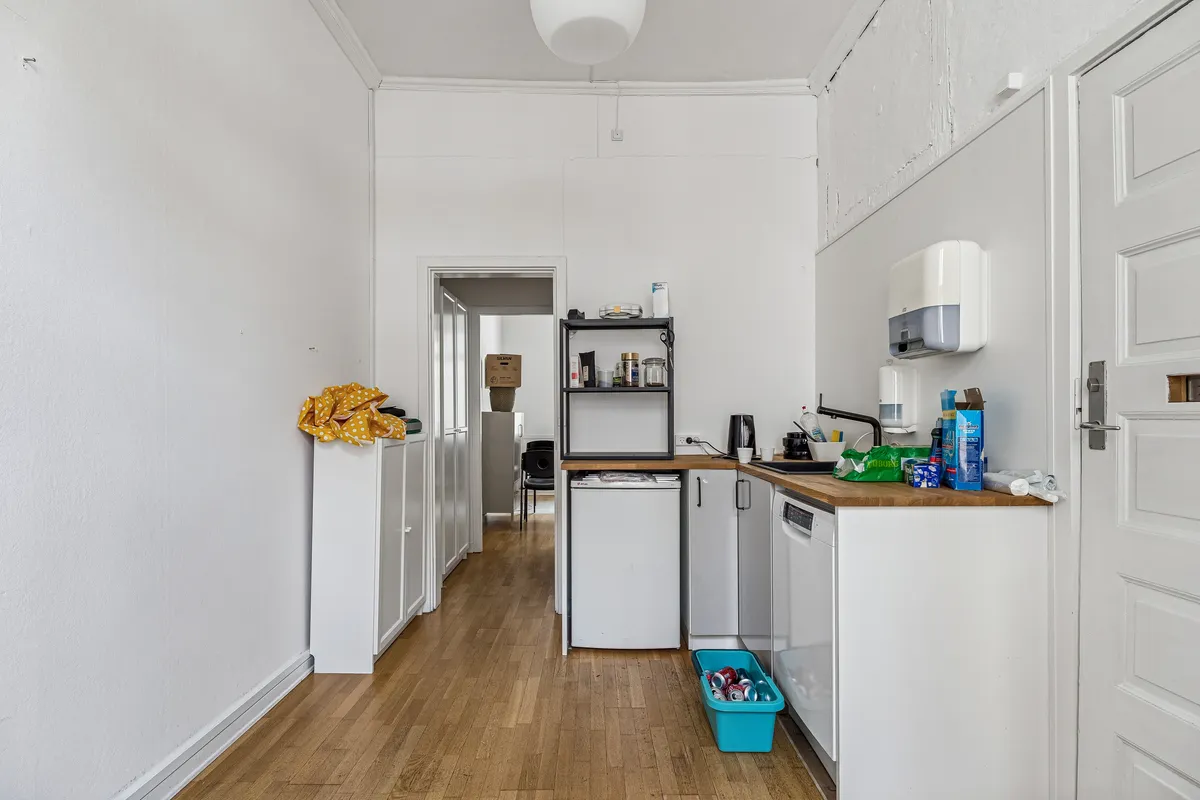 En lys og pænt indrettet køkken med hvide vægge, trægulv, hvide skabe, lysegrå loftslampe og et lille bord med kaffemaskine og andre køkkenartikler.