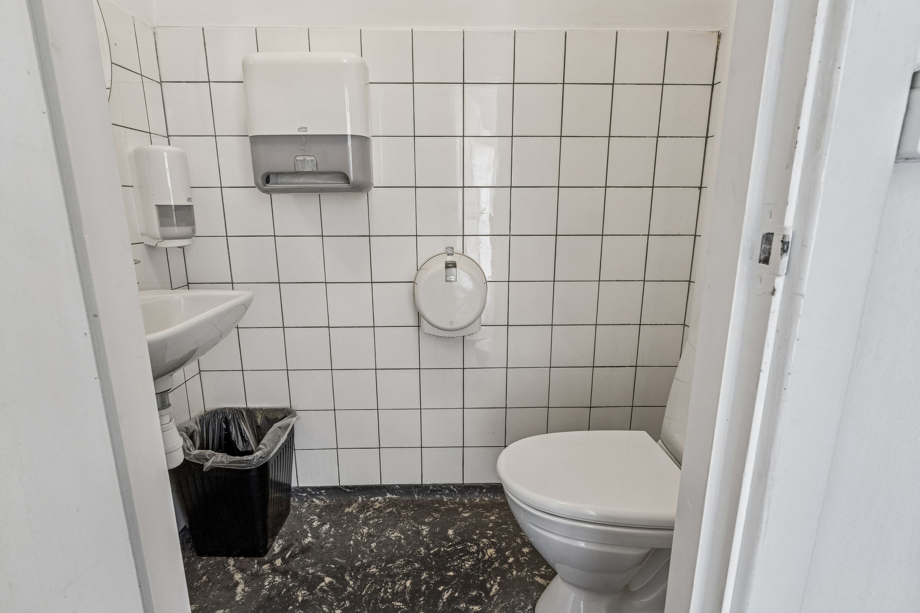 Kontor med toilet og håndvask. Hvidt toilet med håndklædetørrer og sæbeautomat samt sort papkasse. Hvidt toilet og sæbeautomat monteret på væggen