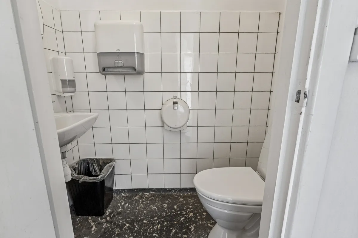 Kontor med toilet og håndvask. Hvidt toilet med håndklædetørrer og sæbeautomat samt sort papkasse. Hvidt toilet og sæbeautomat monteret på væggen