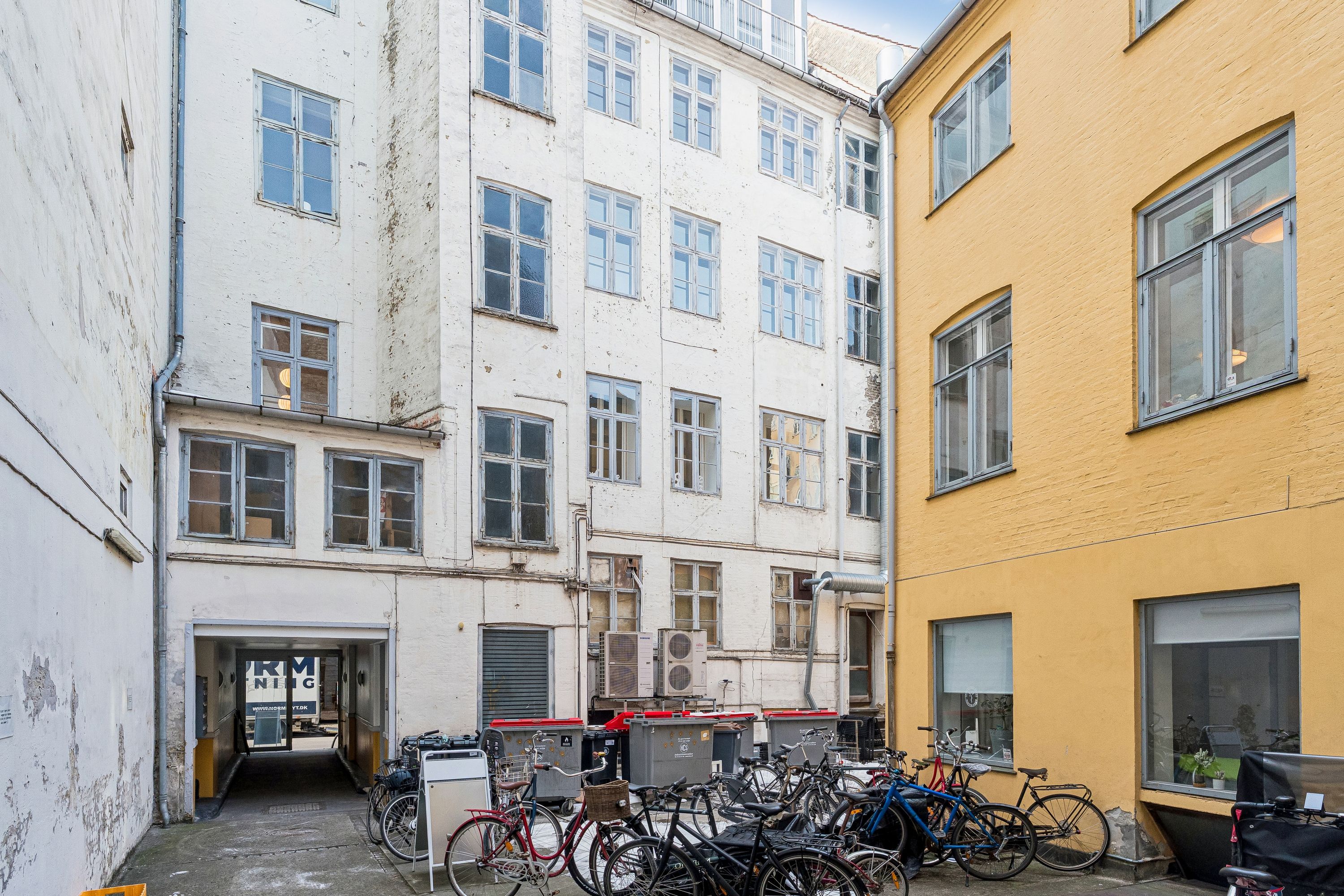 På en gade er der flere lyse og lyse kontorer med mange vinduer. Foran er der parkerede cykler og affaldscontainere. Der er også en dør, der vender ud til en anden gade.