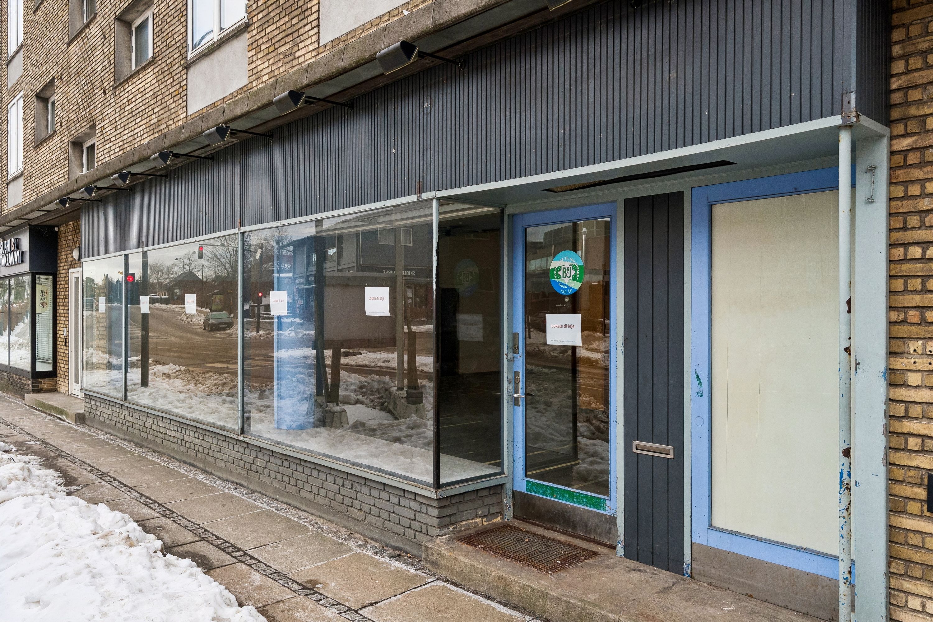 Lejelokale med stor glasfacade og høj synlighed, hjørne af Snogegårdsvej og et tomt gulv med sne. Indgangsdør med postkasse og annoncer, sne dækker fortovet.