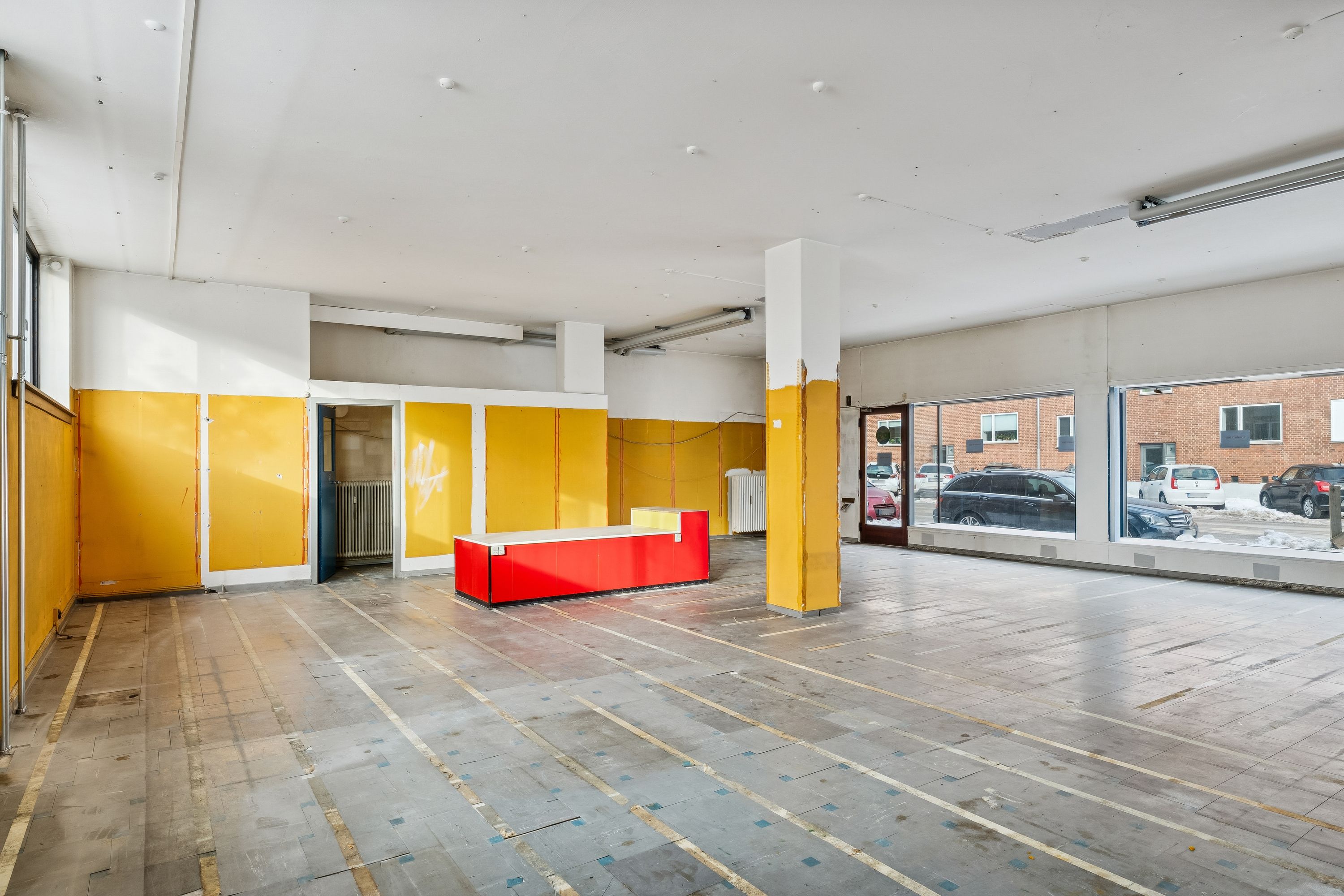 En tom butik med gul og hvid væg og gulv samt to lyse rød hvide skranke og et stort vindue med udsigt til parkeringsplads og biler