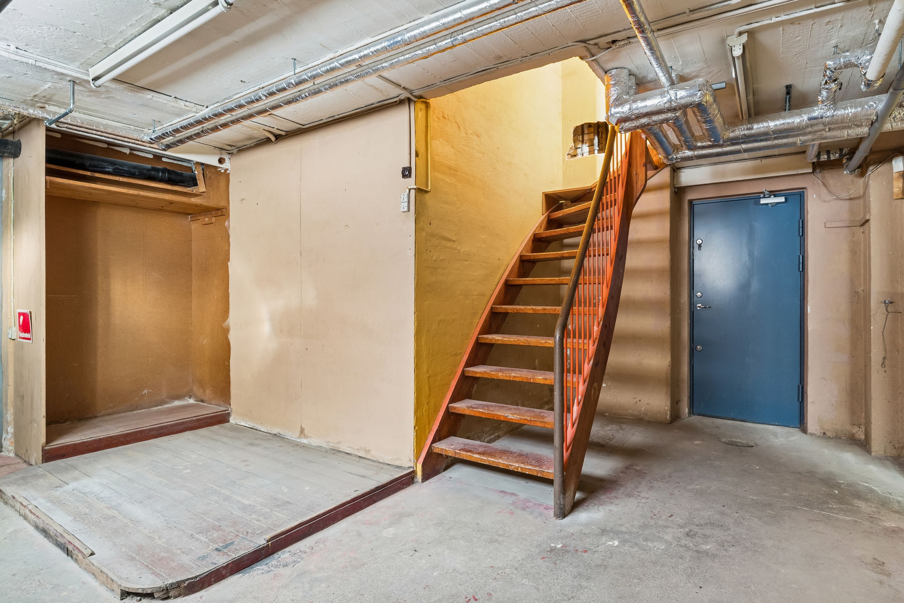 Lokale med trappe og trægulv, åbne døre og røretøj i loftet, en lukket dør med rødt mærke på væggen og en lysstofre, der er tændt
