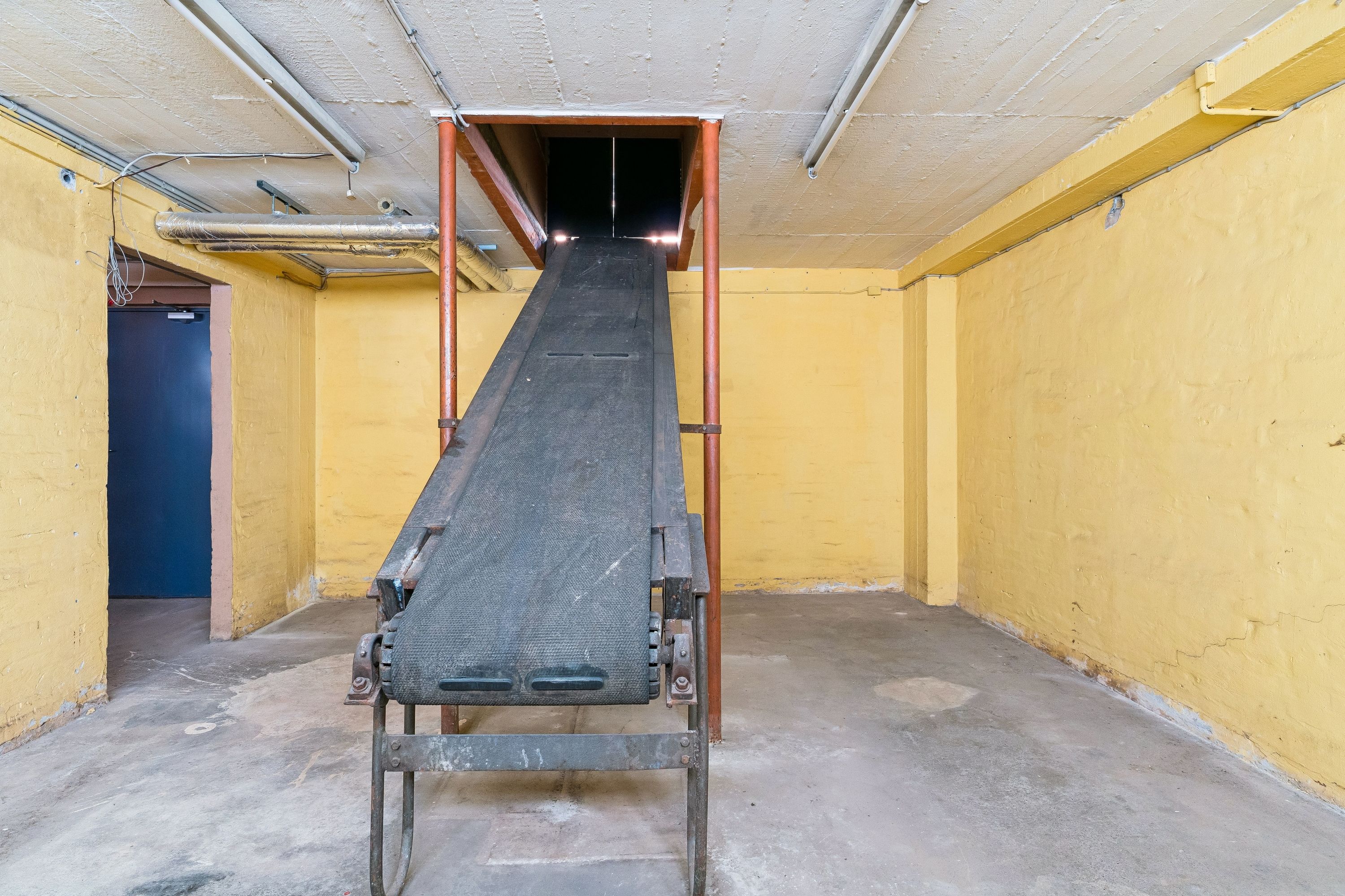 En gammel transportbåndsmaskine står i en gammel kælder med gule vægge og røde søjler. Væggene er beskadiget og der er en åbning i loftet.