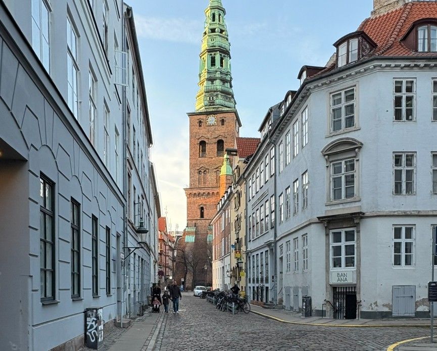 Mennesker vandrer langs en middelalderbygning, der er i flere etager og har et klokketårn, står i et ældre byområde med brostensbelagte gader